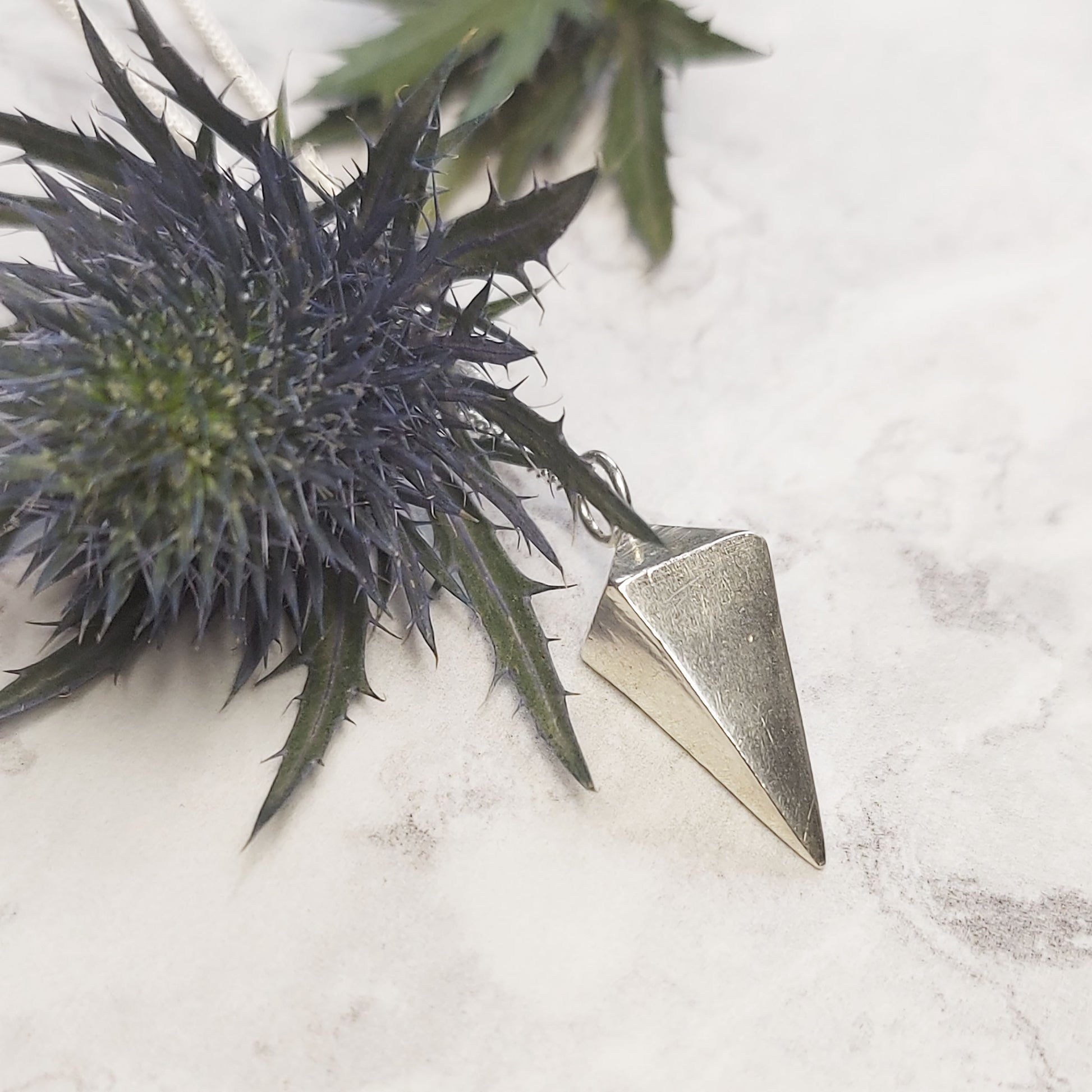 A silver 3D diamond shaped pendant on a silver chain. Pictured with a thistle flower.