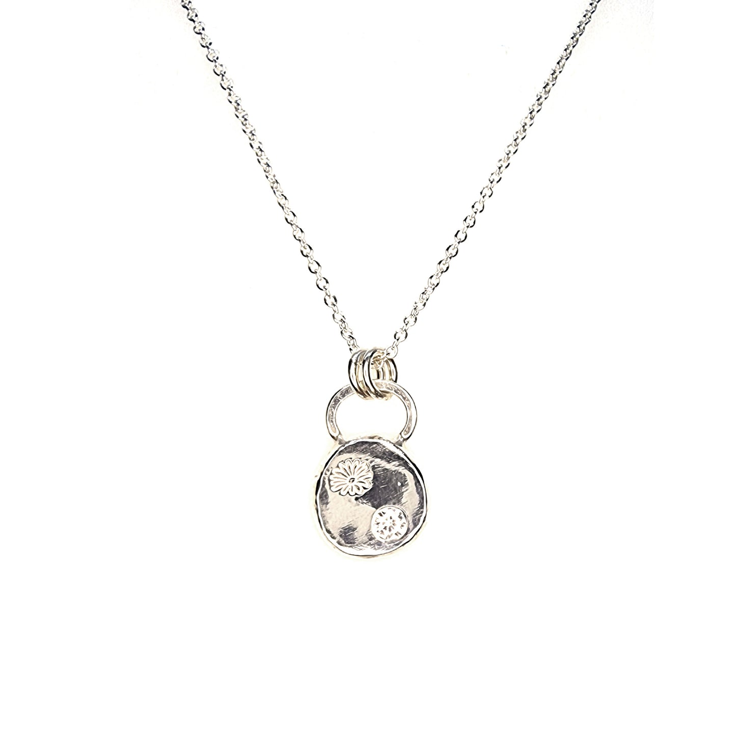A round silver pendant with a daisy flower embossed on it and set with a clear CZ gemstone. On a silver chain.