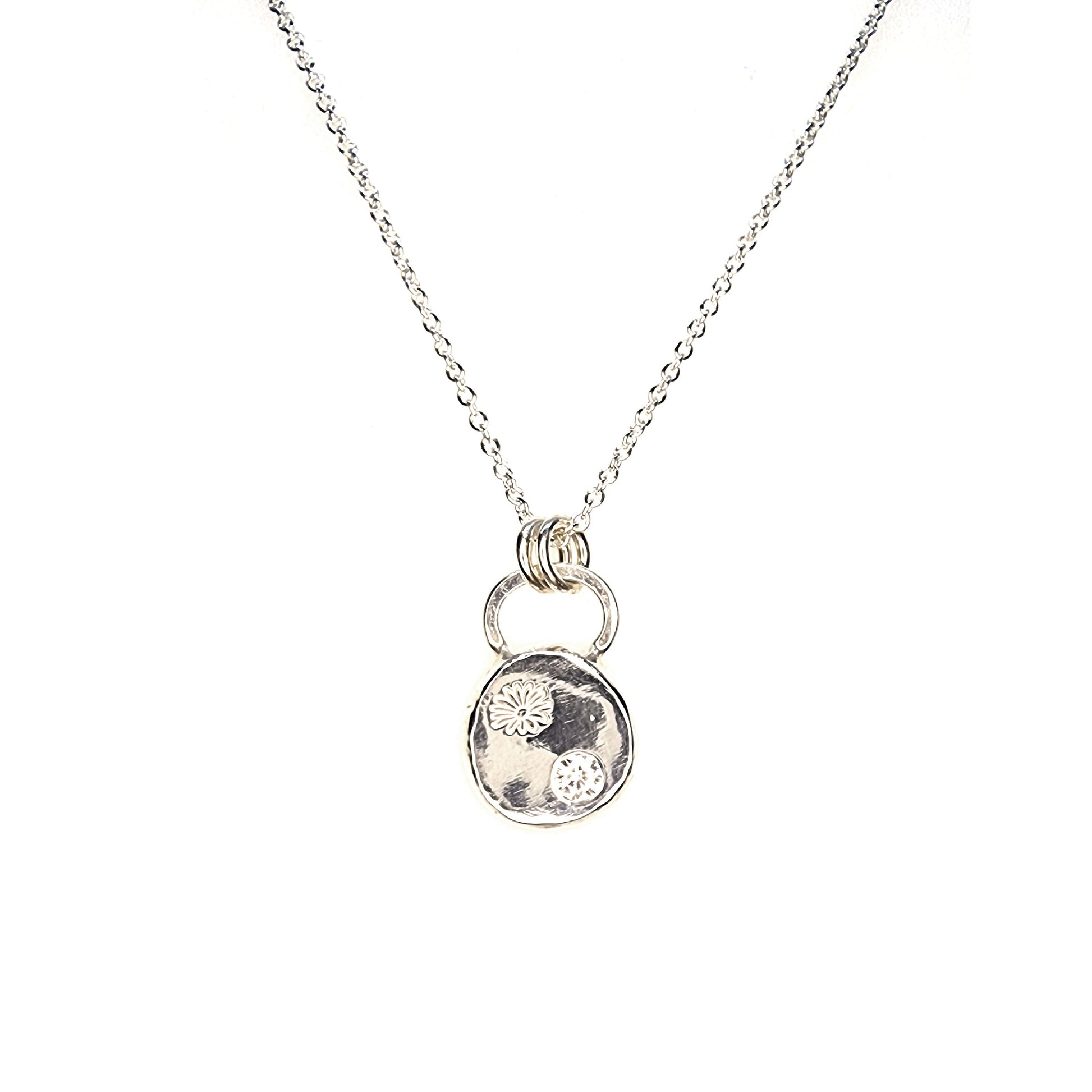 A round silver pendant with a daisy flower embossed on it and set with a clear CZ gemstone. On a silver chain.