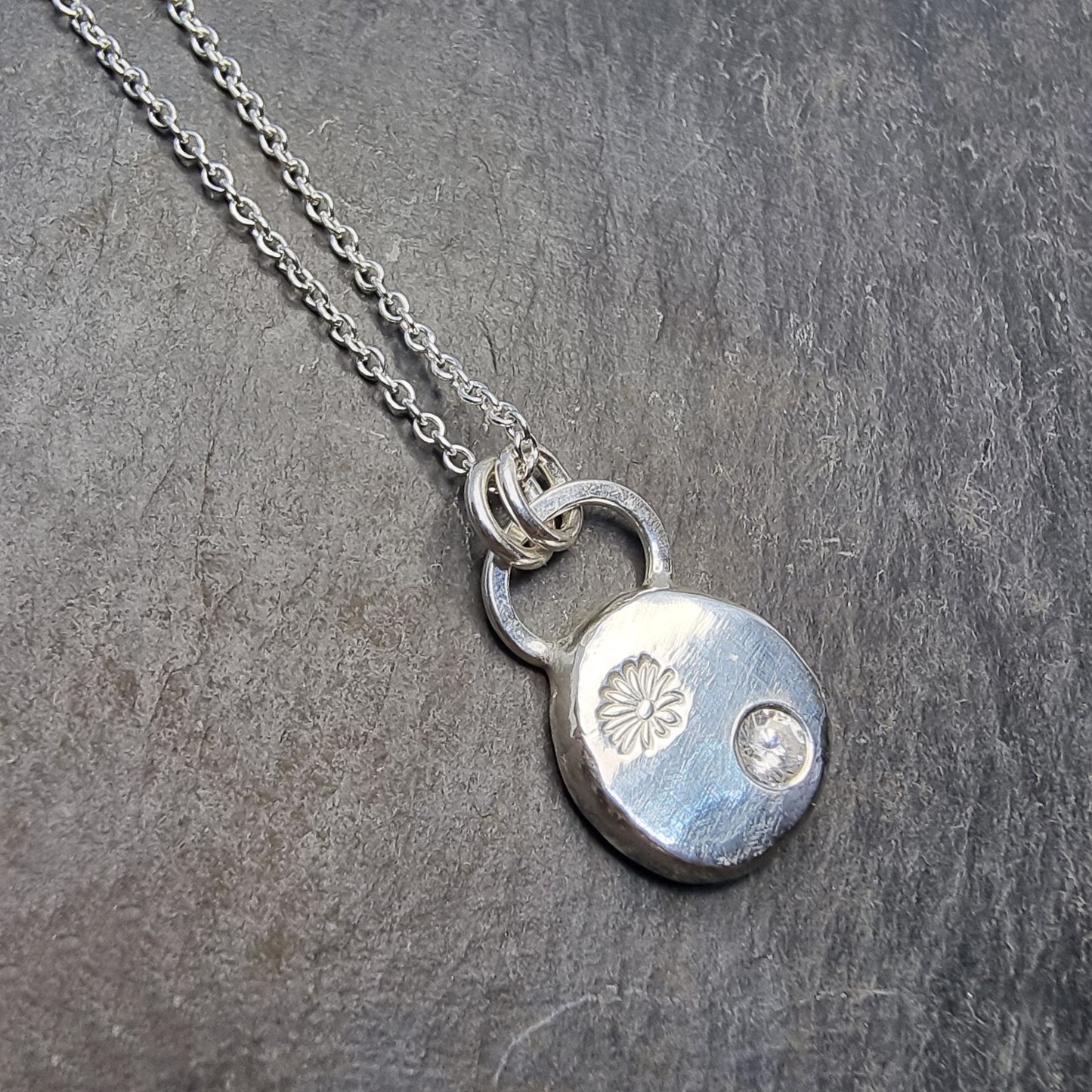 A round silver pendant with a daisy flower embossed on it and set with a clear CZ gemstone. On a silver chain.