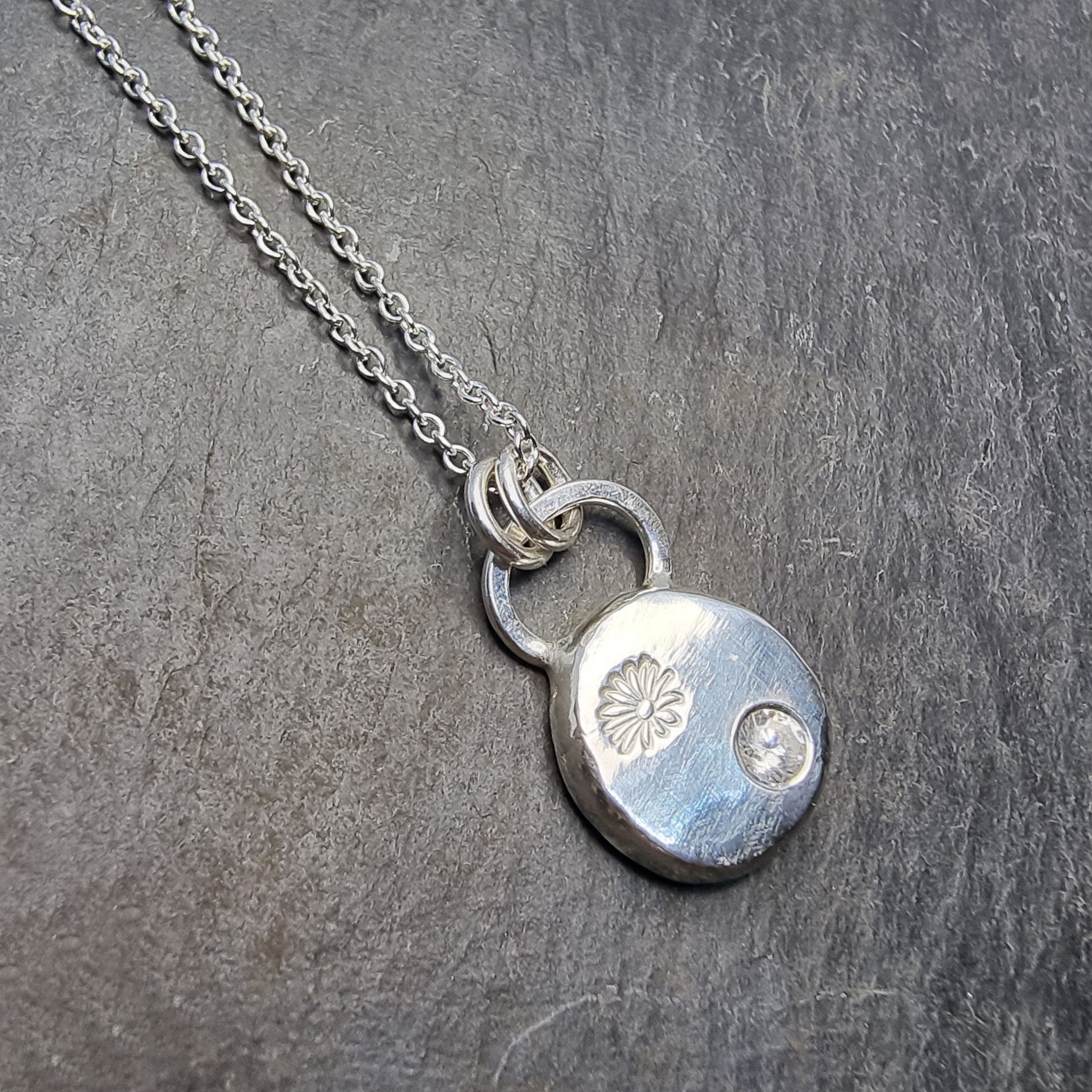 A round silver pendant with a daisy flower embossed on it and set with a clear CZ gemstone. On a silver chain.