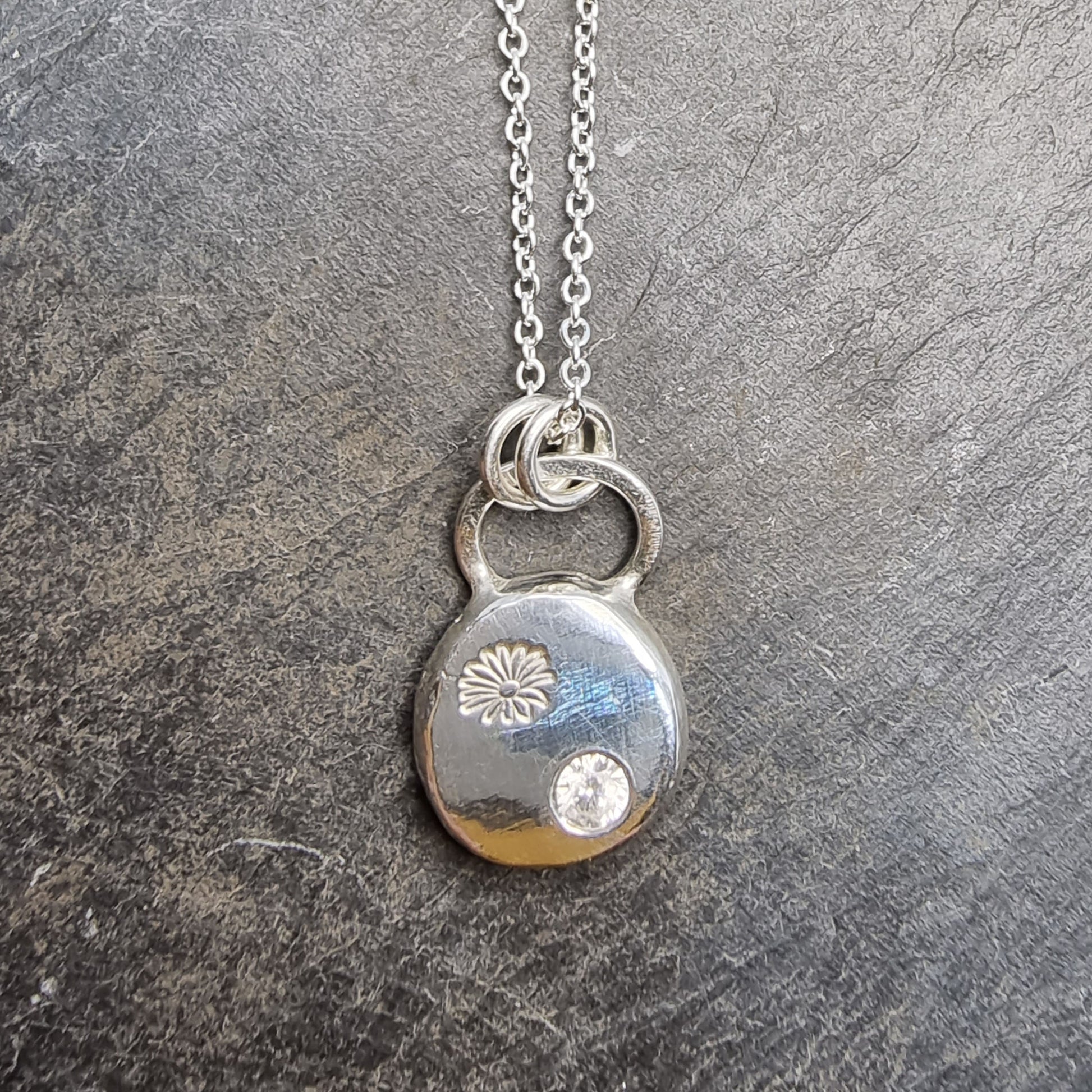 A round silver pendant with a daisy flower embossed on it and set with a clear CZ gemstone. On a silver chain.