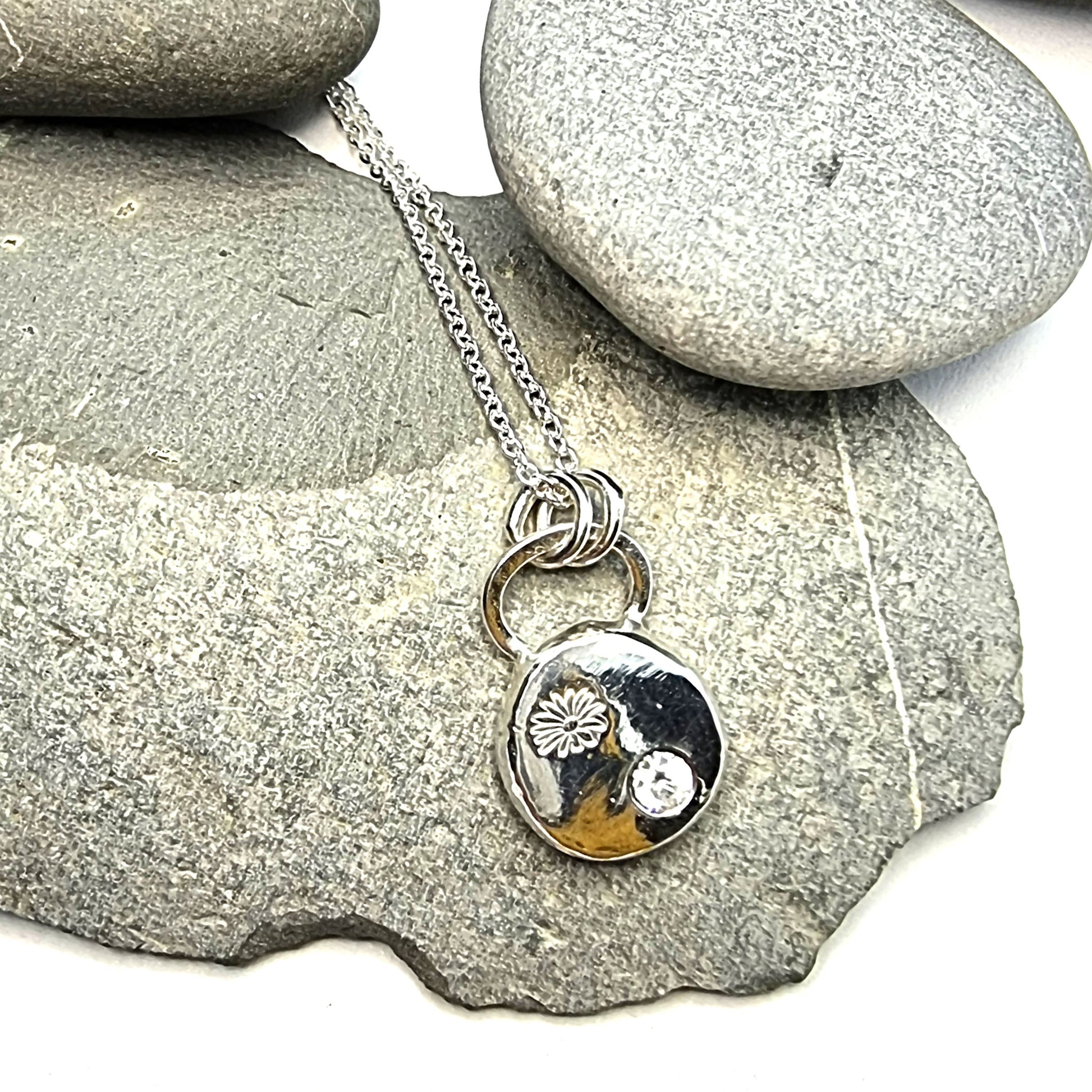 A round silver pendant with a daisy flower embossed on it and set with a clear CZ gemstone. On a silver chain. - on stones.