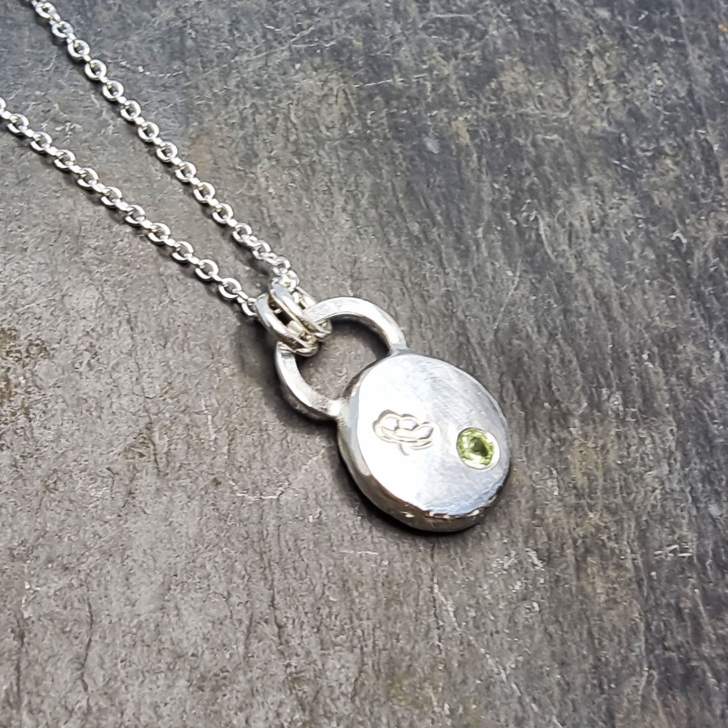 Silver round flat 'pebble' birthstone pendant with a poppy engraved on it and a pale green peridot gemstone on a silver chain. - August
