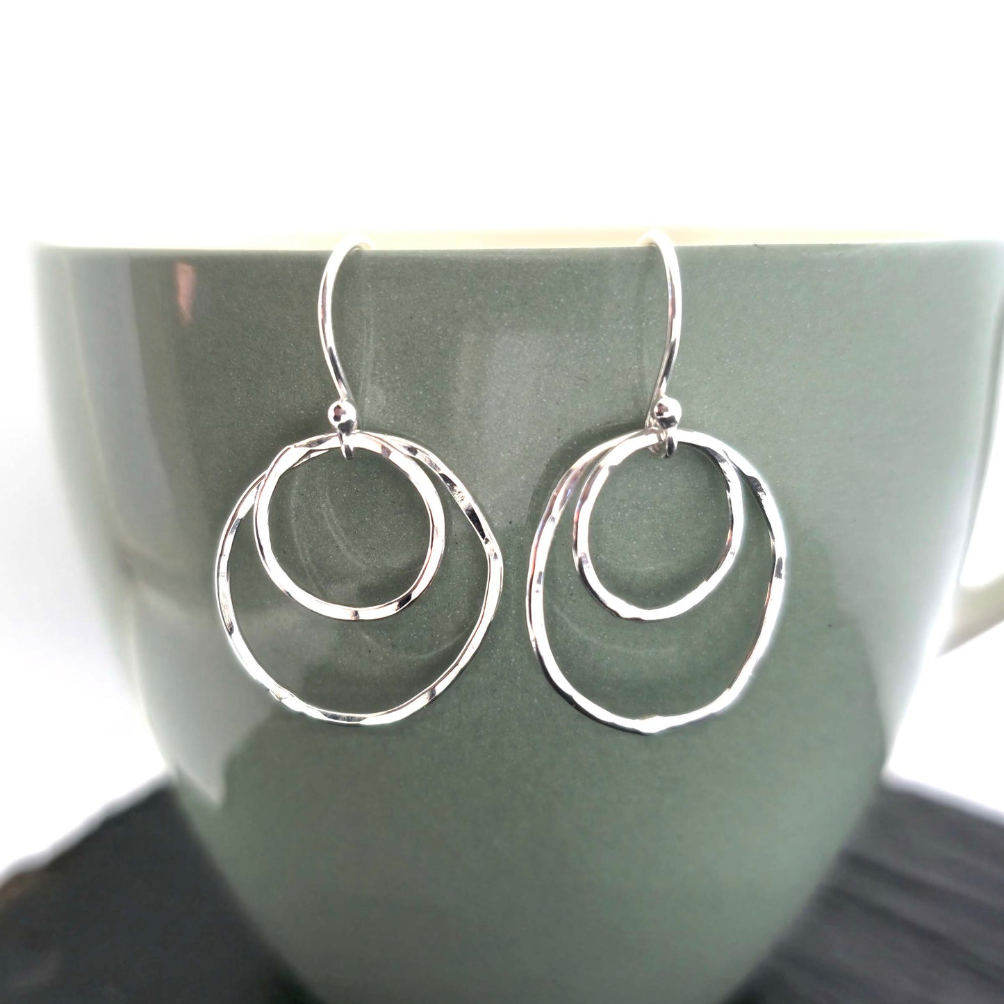 A pair of silver drop earrings. Each earring has 2 rustic circles of different sizes on top of each other with a hammered finish. Displayed on a green cup.