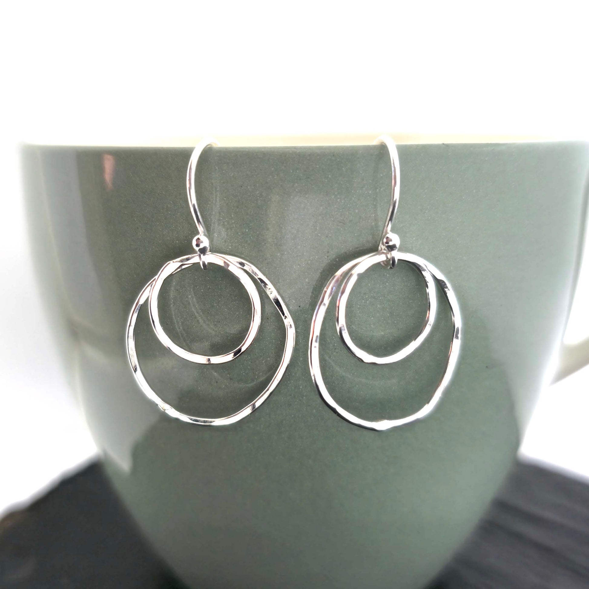 A pair of silver drop earrings. Each earring has 2 rustic circles of different sizes on top of each other with a hammered finish. Displayed on a green cup.