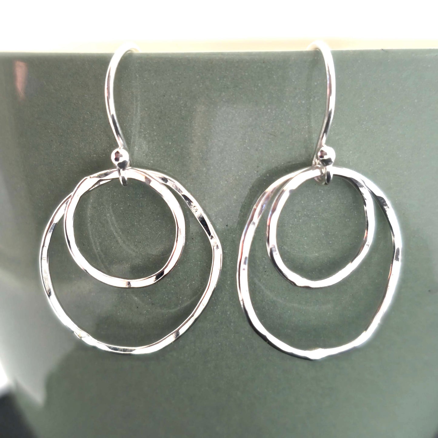 A pair of silver drop earrings. Each earring has 2 rustic circles of different sizes on top of each other with a hammered finish. Displayed on a green cup.