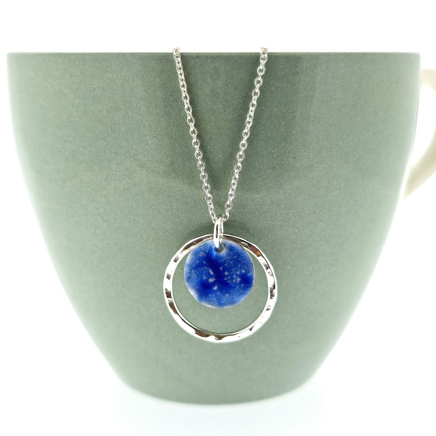 A silver pendant with a central disc with a blue patterned enamel surrounded by a silver hammered open circle. On a silver chain.