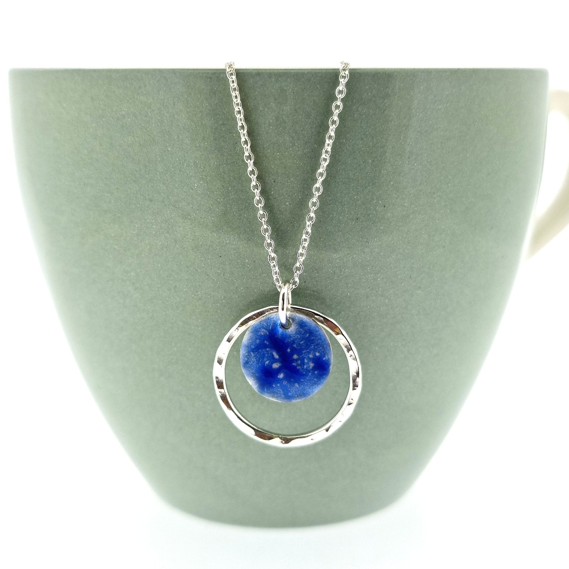 A silver pendant with a central disc with a blue patterned enamel surrounded by a silver hammered open circle. On a silver chain.