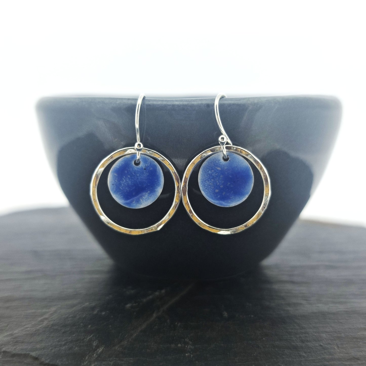 Silver drop earrings with a central disc featuring patterned dark blue enamel surrounded by a silver hammered open circle. Pictured on a dark bowl.
