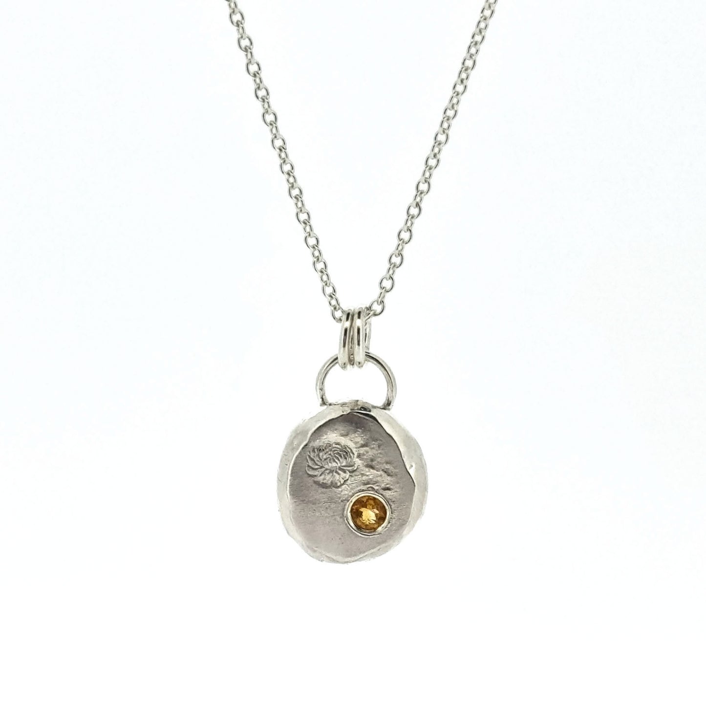 Round silver flat pebble pendant with chrysanthemum flower engraved on it and a flush set yellow citrine gemstone. On a silver chain.