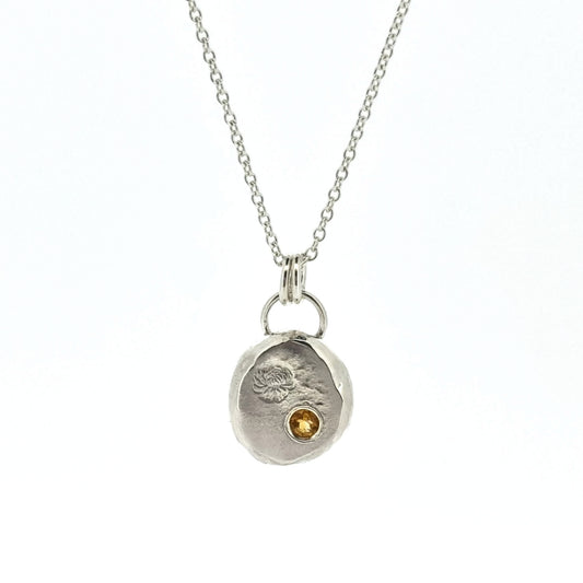 Round silver flat pebble pendant with chrysanthemum flower engraved on it and a flush set yellow citrine gemstone. On a silver chain.