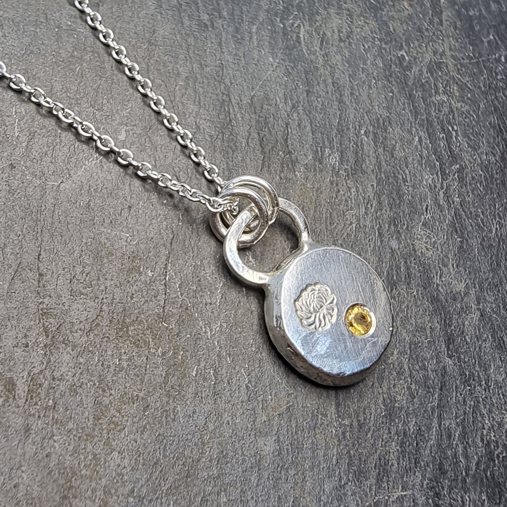 Round silver flat pebble pendant with chrysanthemum flower engraved on it and a flush set yellow citrine gemstone. On a silver chain.
