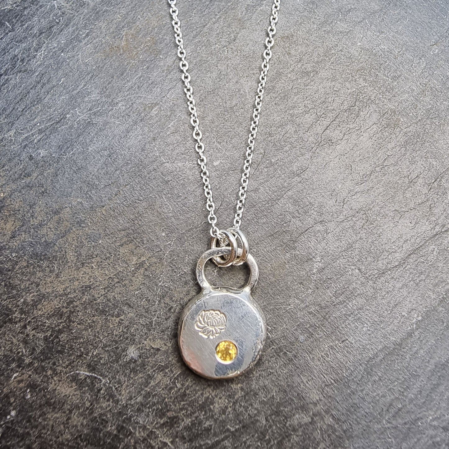 Round silver flat pebble pendant with chrysanthemum flower engraved on it and a flush set yellow citrine gemstone. On a silver chain.