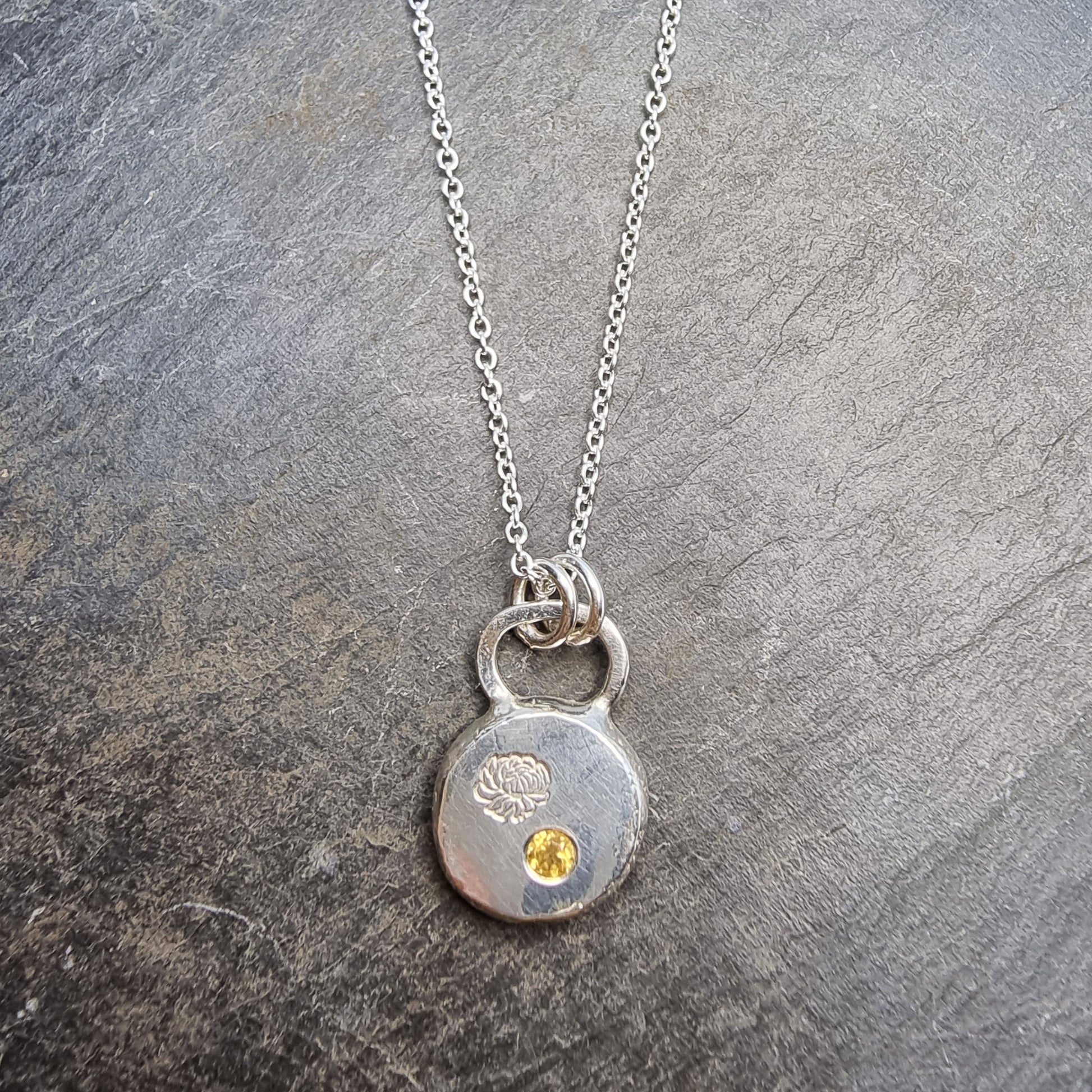 Round silver flat pebble pendant with chrysanthemum flower engraved on it and a flush set yellow citrine gemstone. On a silver chain.