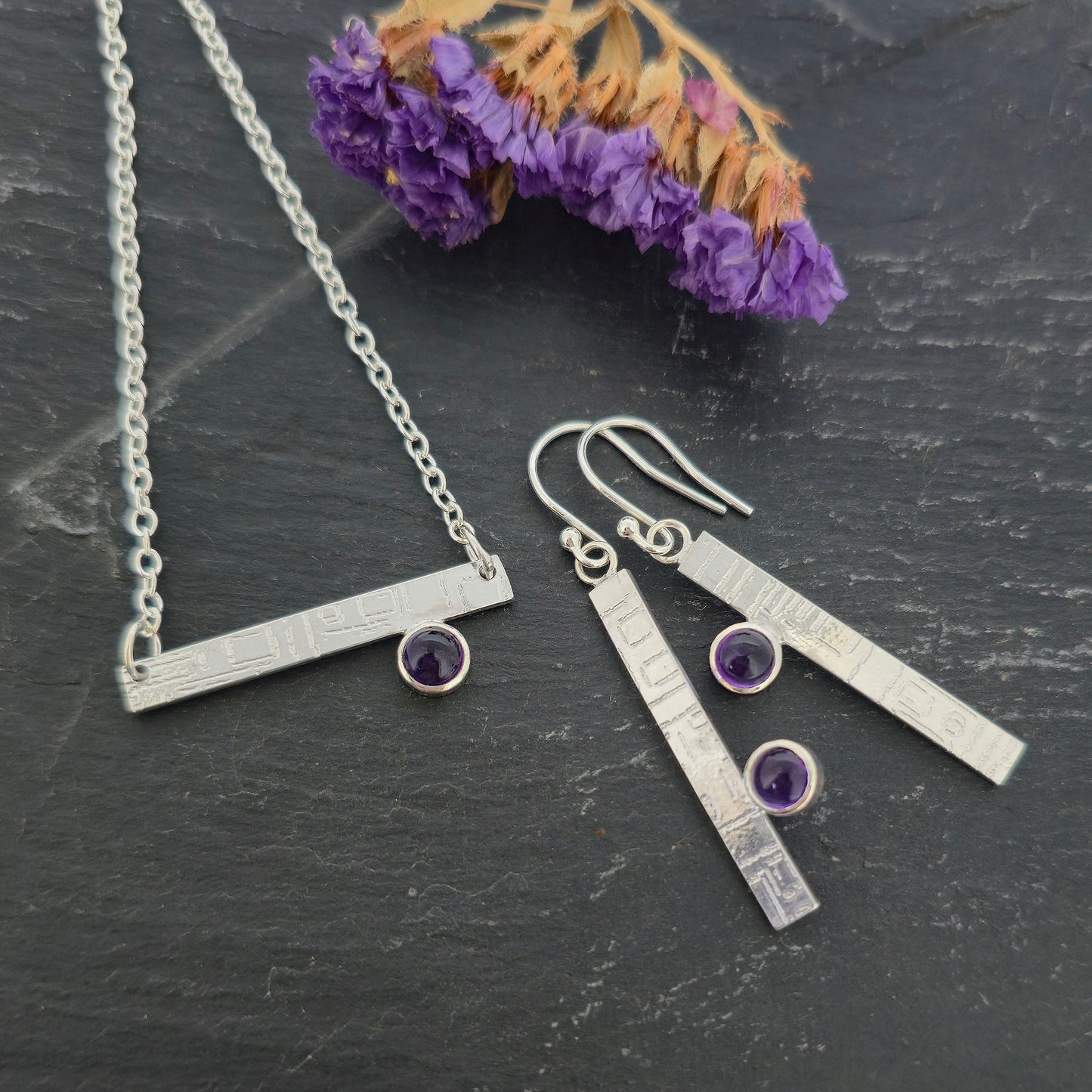 Handmade silver jewellery set including a bar necklace and matching drop earrings with small purple amethyst gemstones, displayed on a dark textured surface with dried purple flowers. Mijoux Jewellery.
