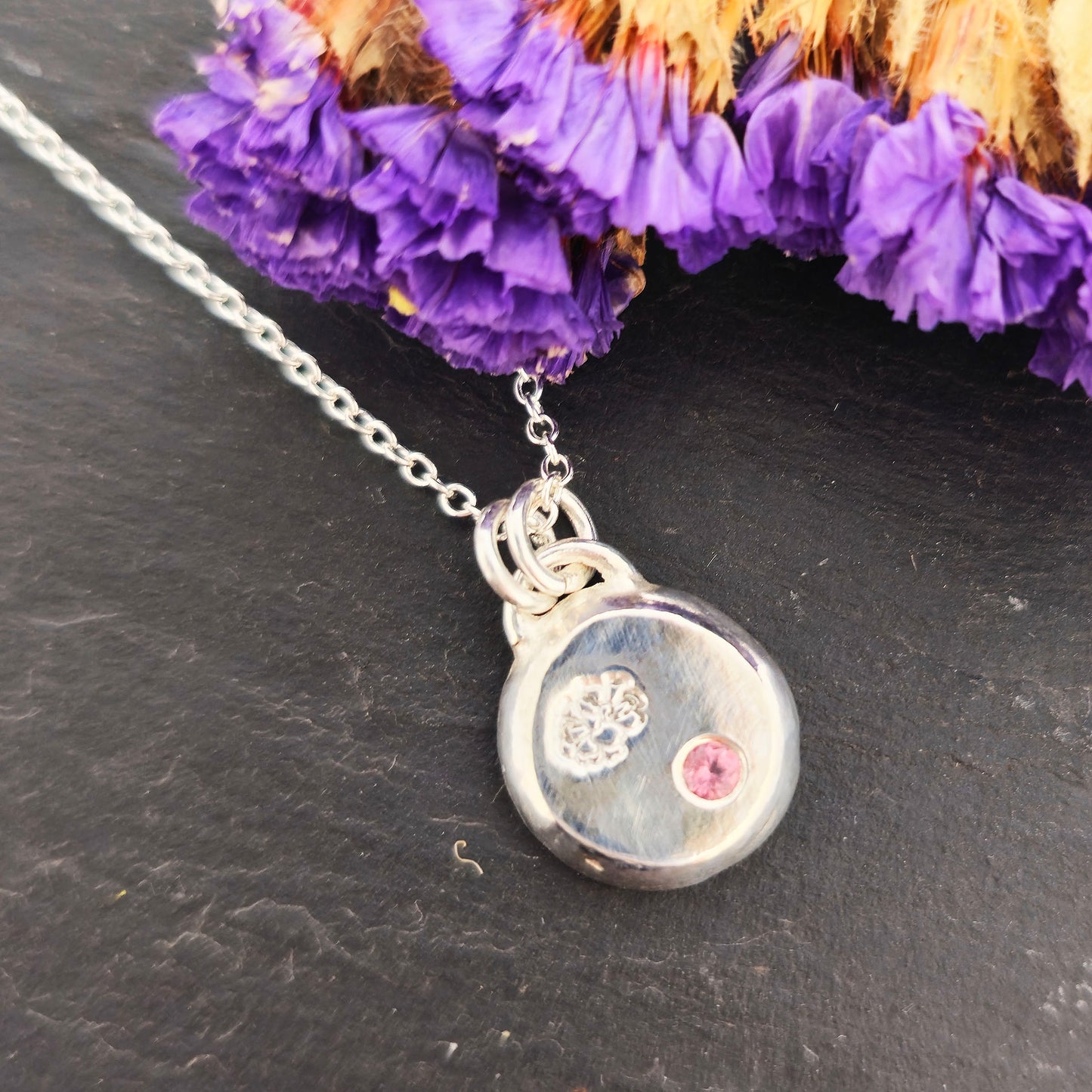 Round silver pendant with an engraved marigold flower and a flush set pink tourmaline gemstone. On a silver chain. On a black background with dried flowers.
