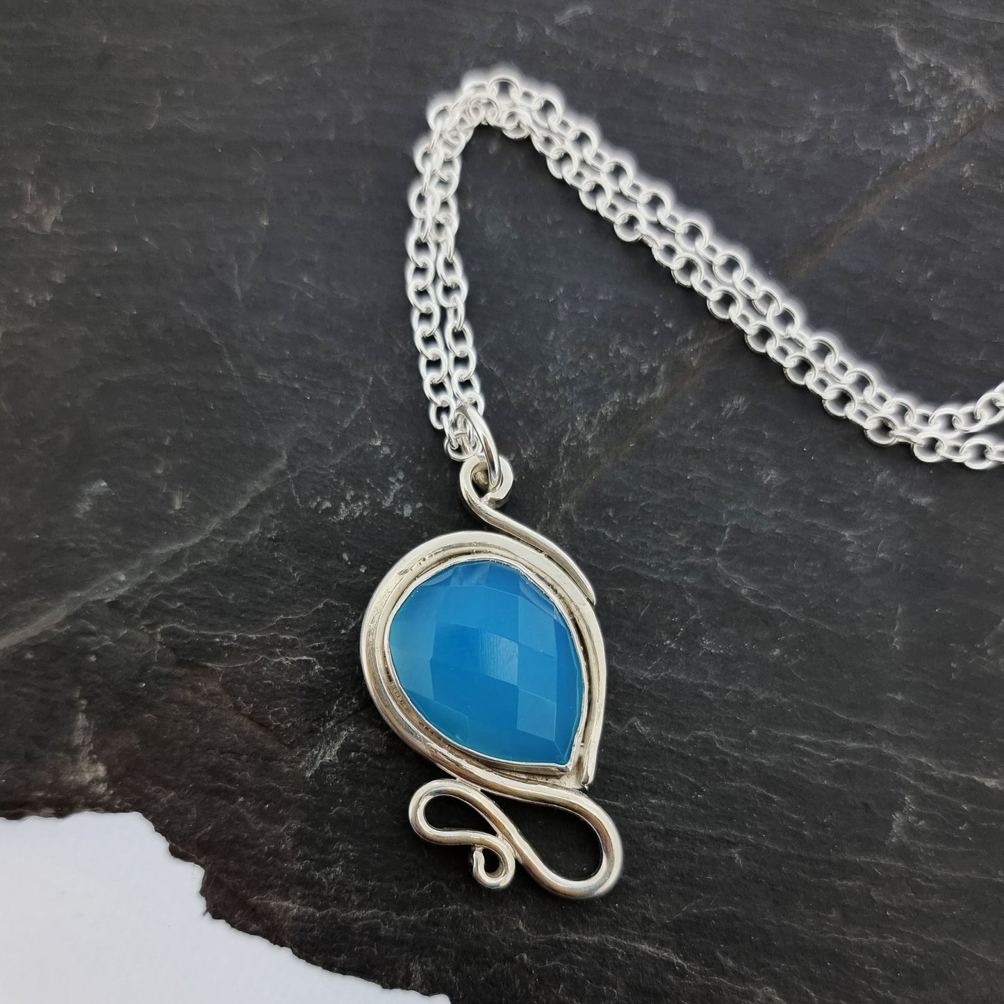 Silver pendant necklace with a teardrop shaped turquoise blue chalcedony gemstone surrounded by a silver bezel and with silver swirls above and below. On a silver chain.