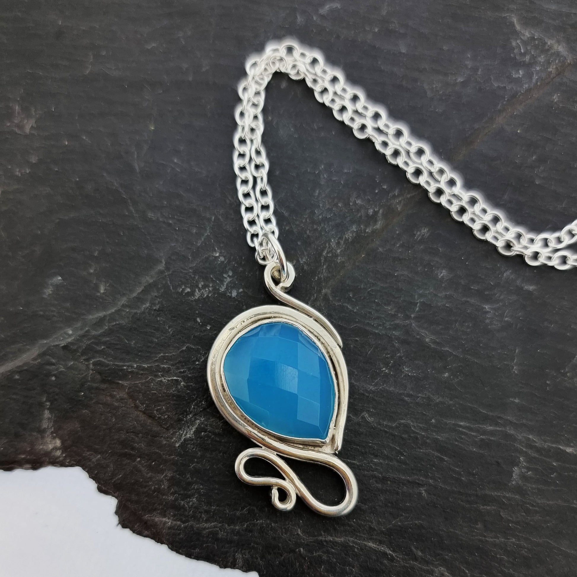 Silver pendant necklace with a teardrop shaped turquoise blue chalcedony gemstone surrounded by a silver bezel and with silver swirls above and below. On a silver chain.