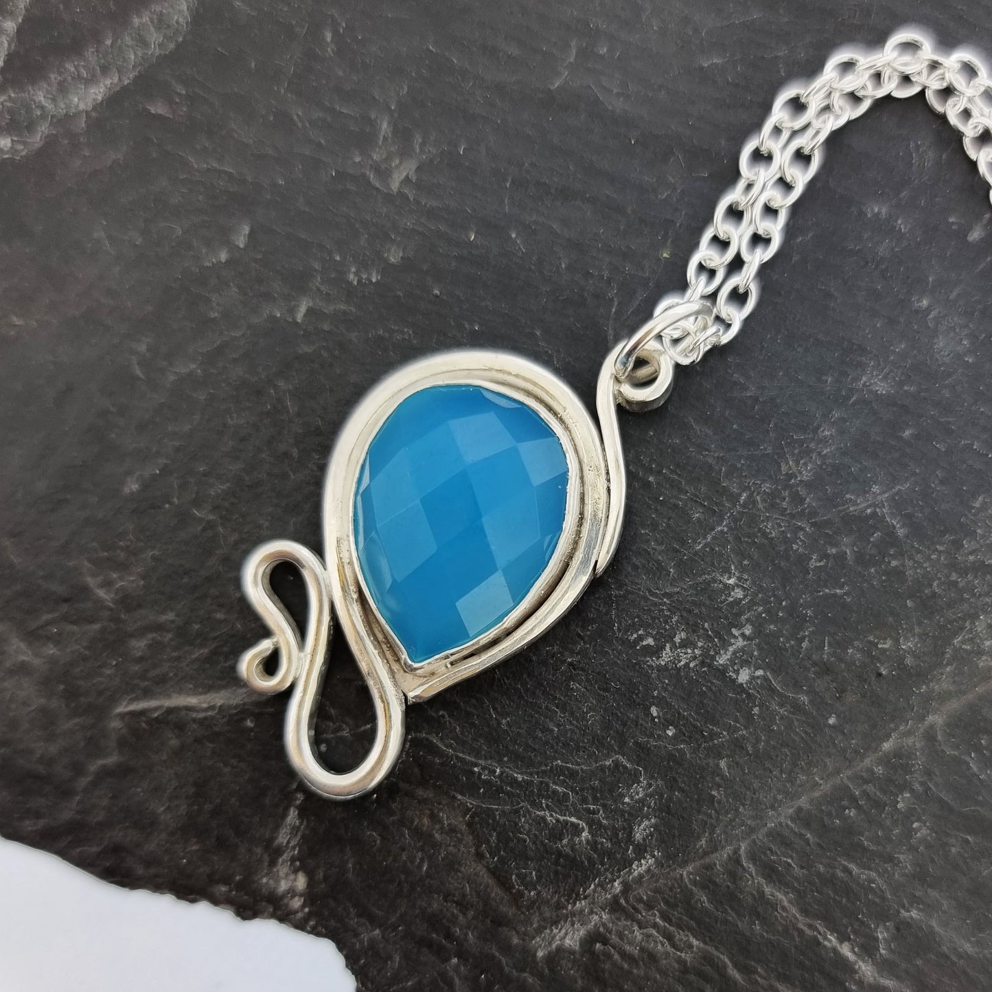 Silver pendant necklace with a teardrop shaped turquoise blue chalcedony gemstone surrounded by a silver bezel and with silver swirls above and below. On a silver chain.