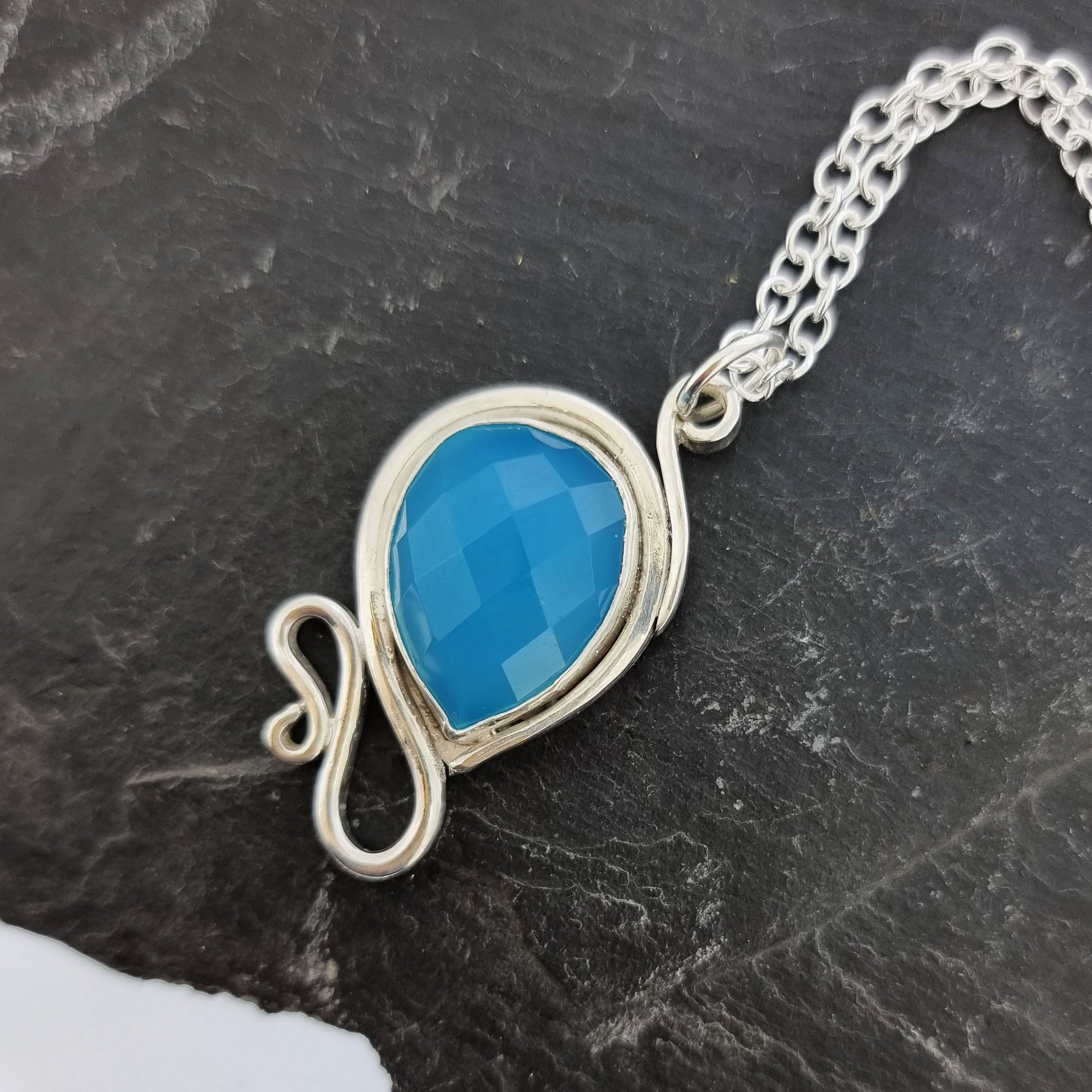 Silver pendant necklace with a teardrop shaped turquoise blue chalcedony gemstone surrounded by a silver bezel and with silver swirls above and below. On a silver chain.