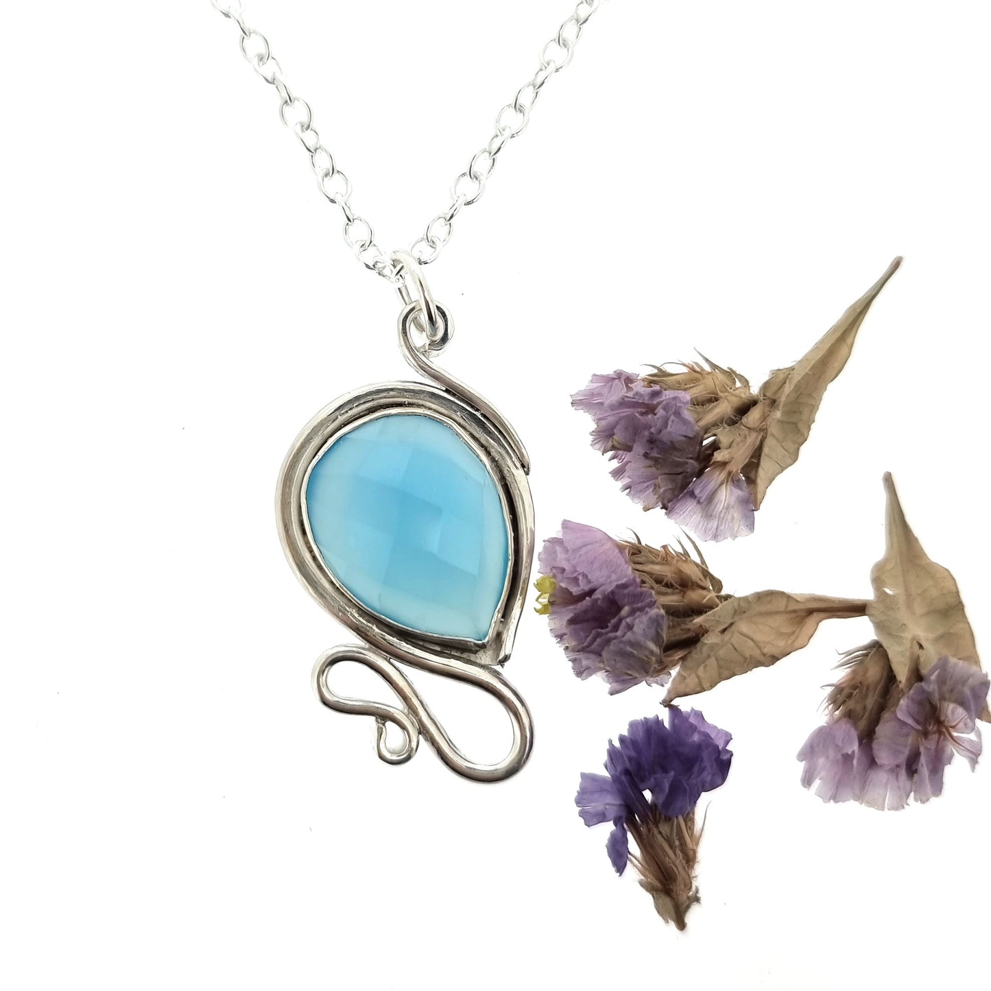Silver pendant necklace with a teardrop shaped turquoise blue chalcedony gemstone surrounded by a silver bezel and with silver swirls above and below. On a silver chain. Pictured with flowers.