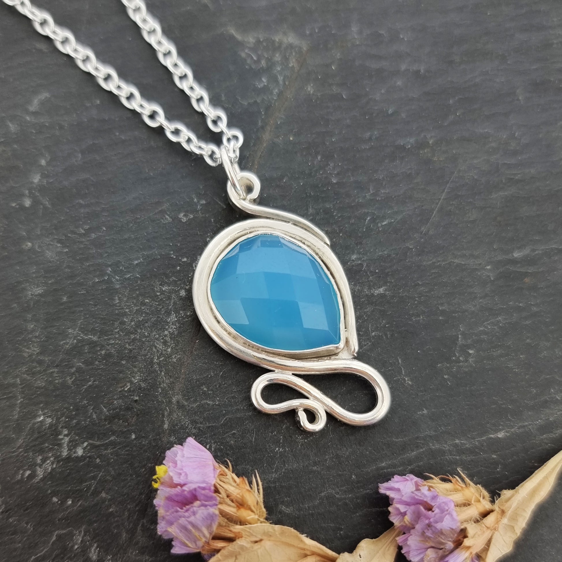 Silver pendant necklace with a teardrop shaped turquoise blue chalcedony gemstone surrounded by a silver bezel and with silver swirls above and below. On a silver chain. Pictured with flowers.