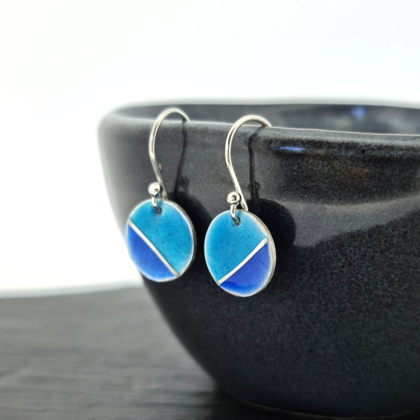 Silver round drop earrings with blue enamels. A silver line runs at an angle across the circle with light turquoise blue in the top part and dark royal blue in the bottom part. Pictured on a bowl.