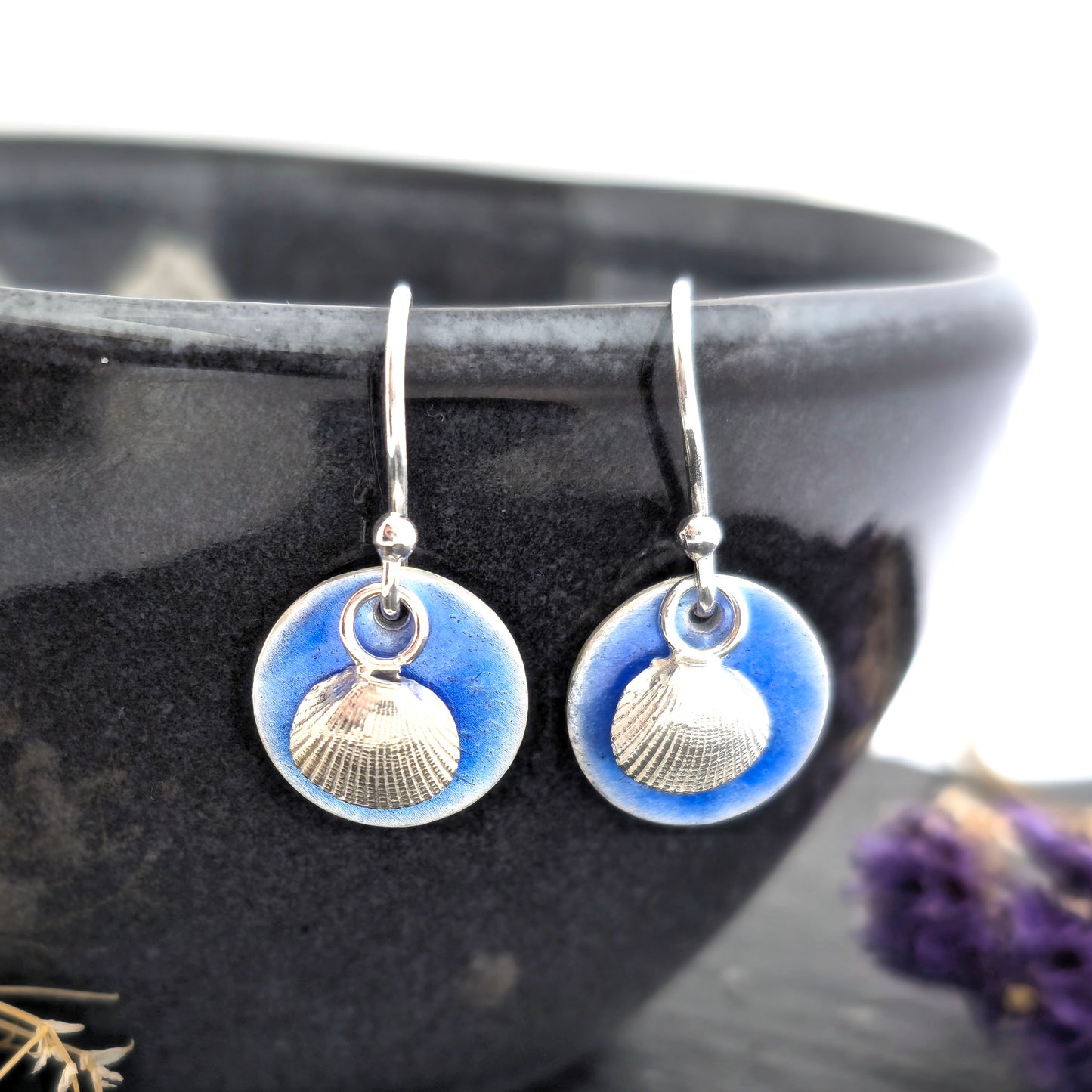 Pair of silver earrings with a blue enamel disc and a silver cockle shell on a black bowl. Handmade. Mijoux Jewellery.