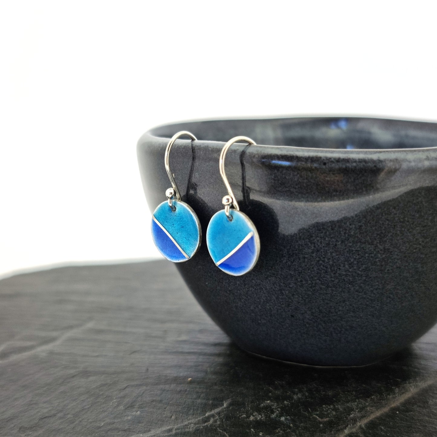 Silver round drop earrings with blue enamels. A silver line runs at an angle across the circle with light turquoise blue in the top part and dark royal blue in the bottom part. Pictured on a bowl.