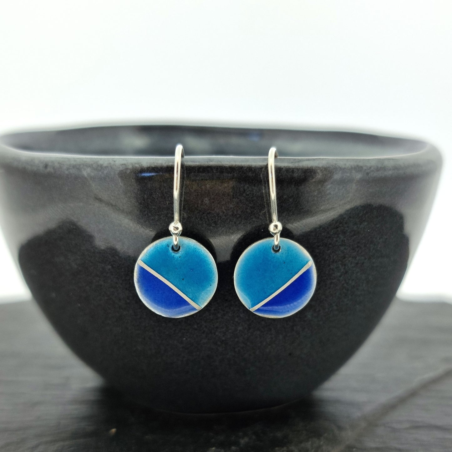 Silver round drop earrings with blue enamels. A silver line runs at an angle across the circle with light turquoise blue in the top part and dark royal blue in the bottom part. Pictured on a bowl.