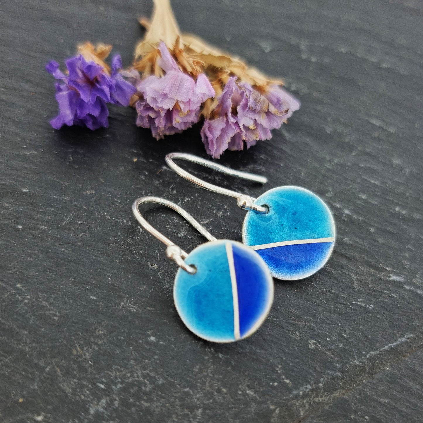 Silver round drop earrings with blue enamels. A silver line runs at an angle across the circle with light turquoise blue in the top part and dark royal blue in the bottom part. Pictured with flowers.