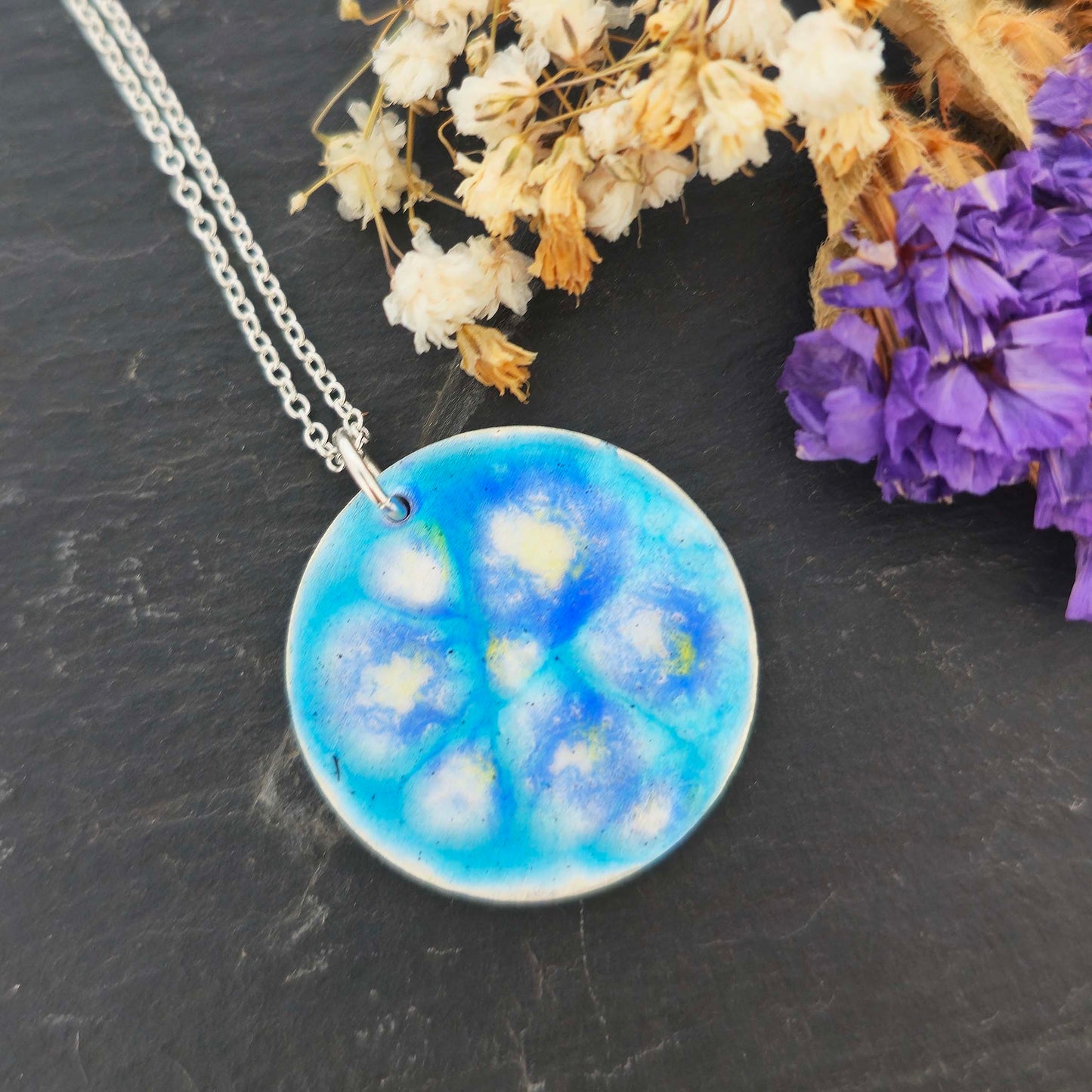 Round silver and blue enamel flower pendant necklace on a dark surface with dried flowers