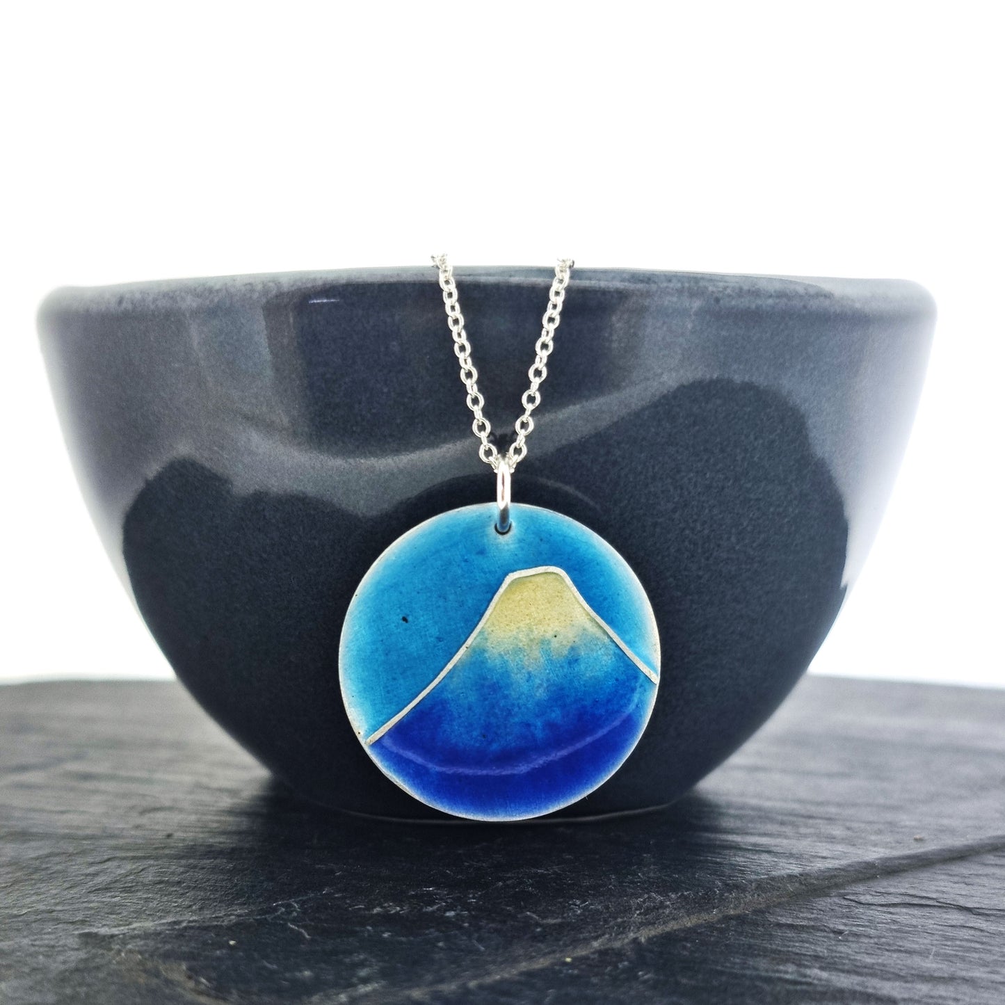 A round silver pendant with a mountain on in blue and white enamels. Suspended on a silver chain. Shown on a black bowl.