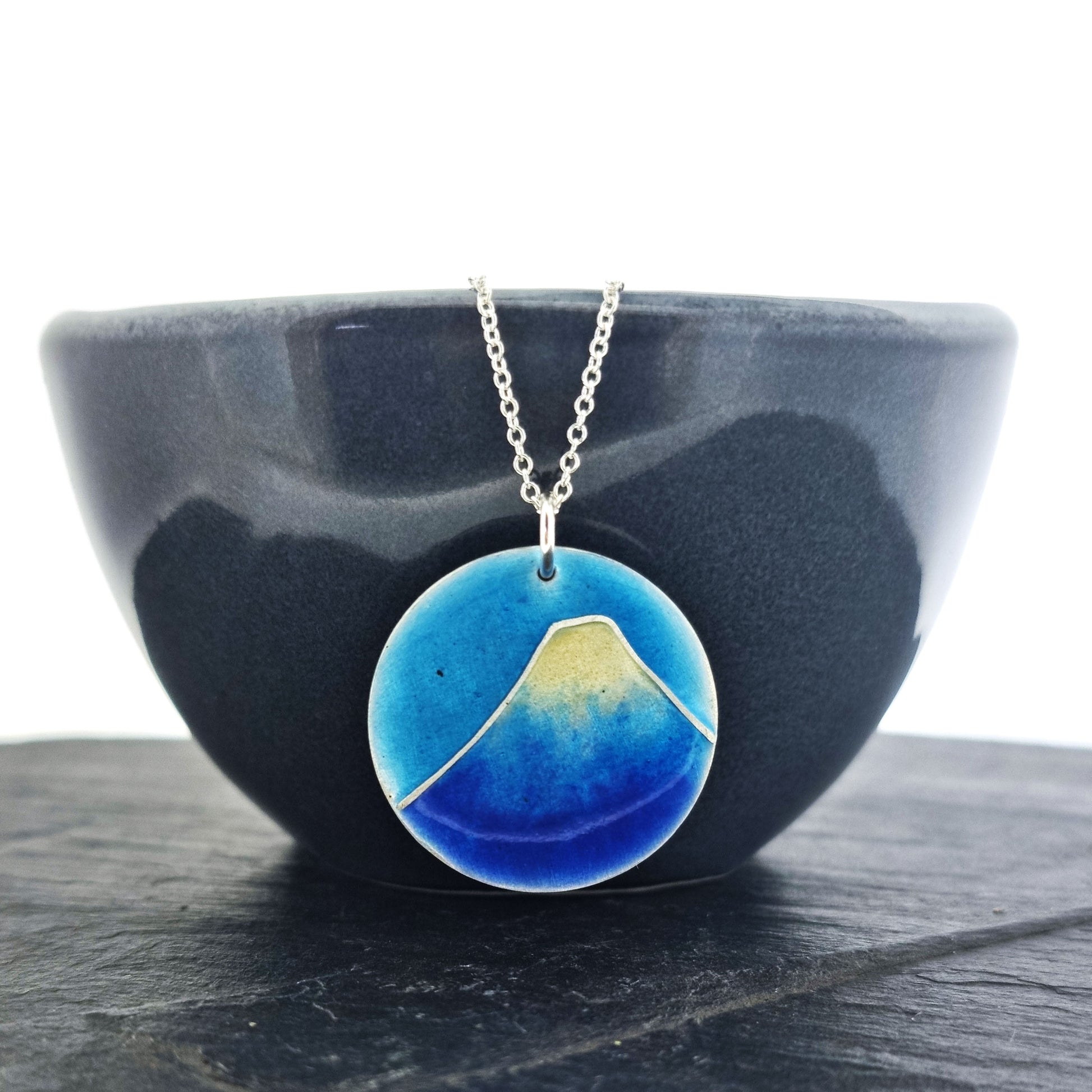 A round silver pendant with a mountain on in blue and white enamels. Suspended on a silver chain. Shown on a black bowl.