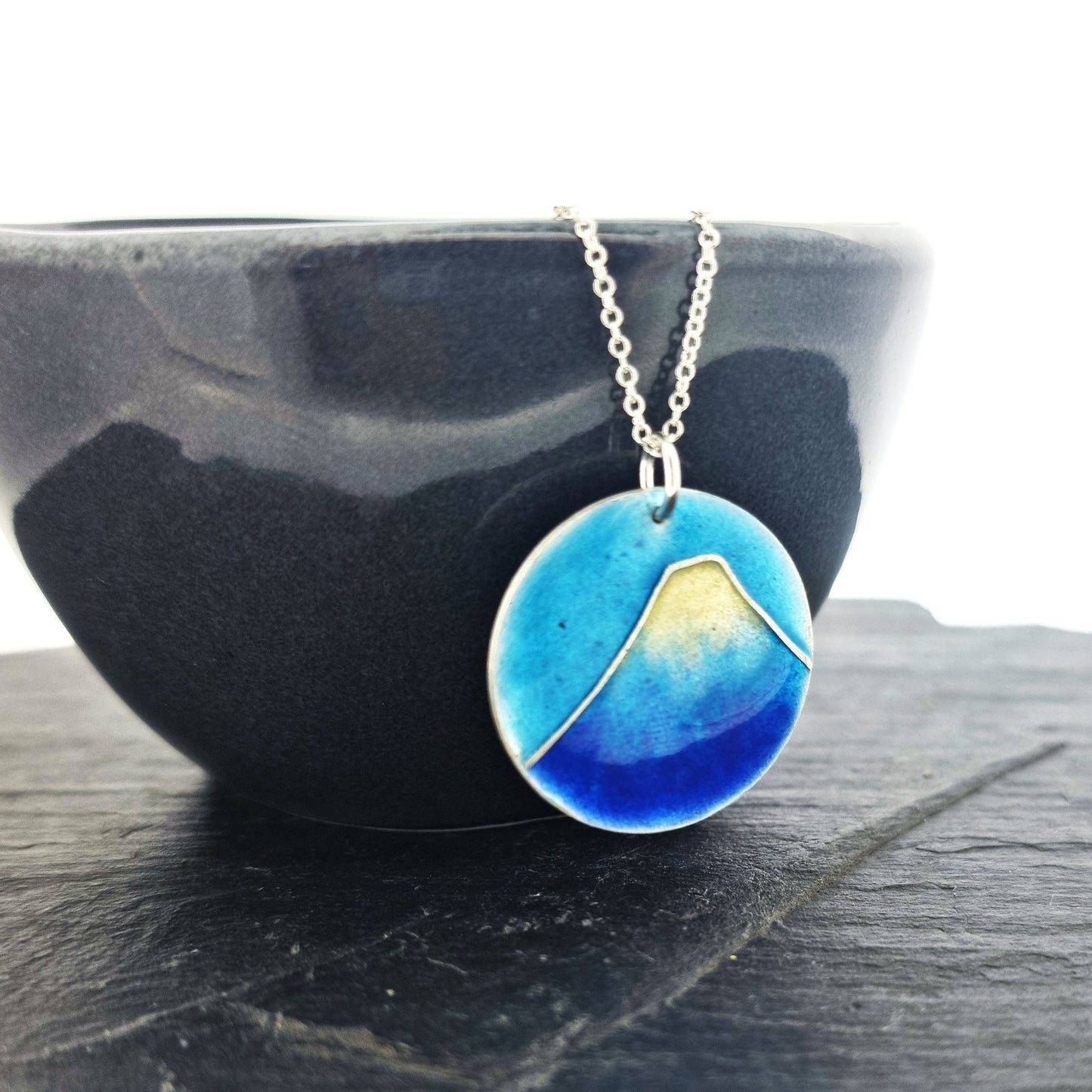 A round silver pendant with a mountain on in blue and white enamels. Suspended on a silver chain. Shown on a black bowl.