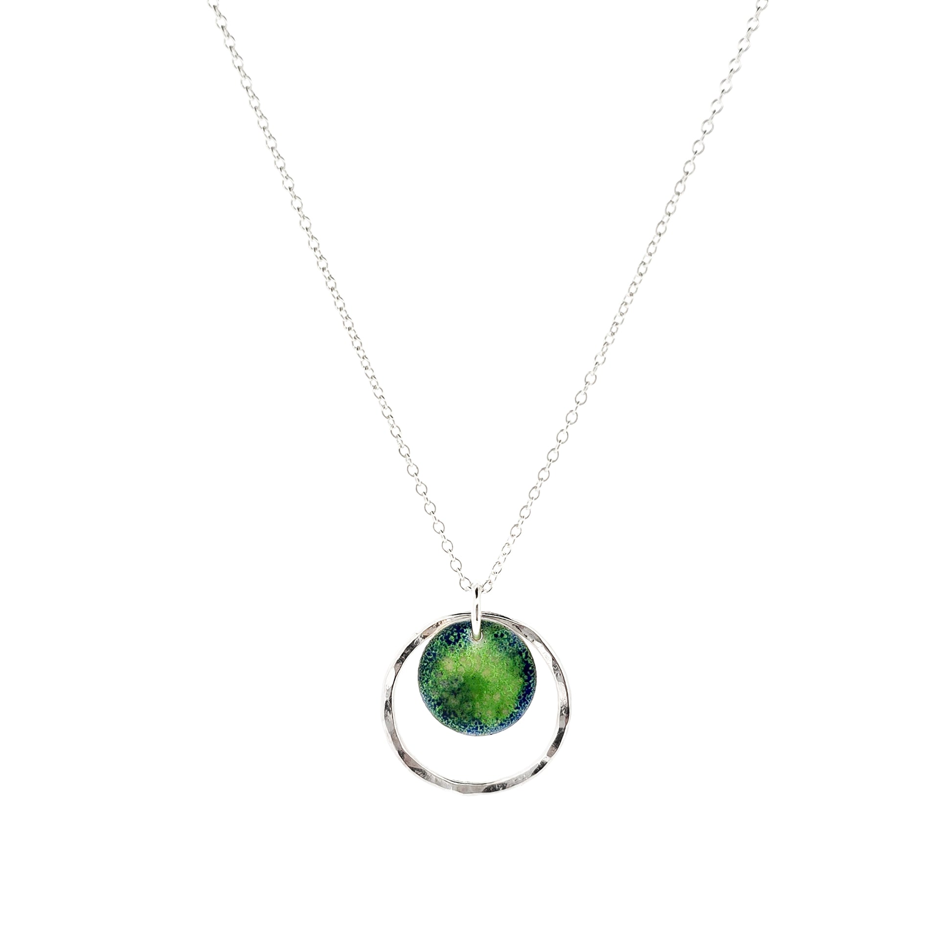 A silver pendant necklace featuring a silver disc with a pattern of green and blue enamel surrounded by a silver circle with a hammered finish. On a silver chain.