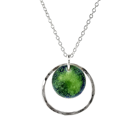 A silver pendant necklace featuring a silver disc with a pattern of green and blue enamel surrounded by a silver circle with a hammered finish. On a silver chain.