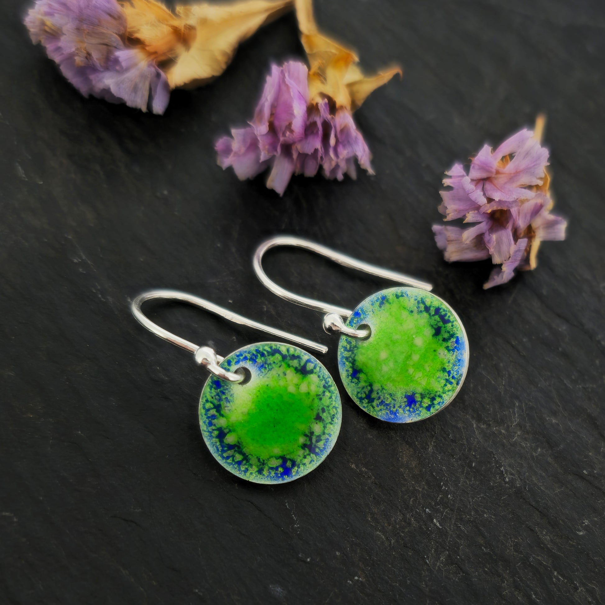 Silver earrings featuring a disc coloured with a pattern of green and blue enamel on silver ear hooks. Pictured on slate with flowers.
