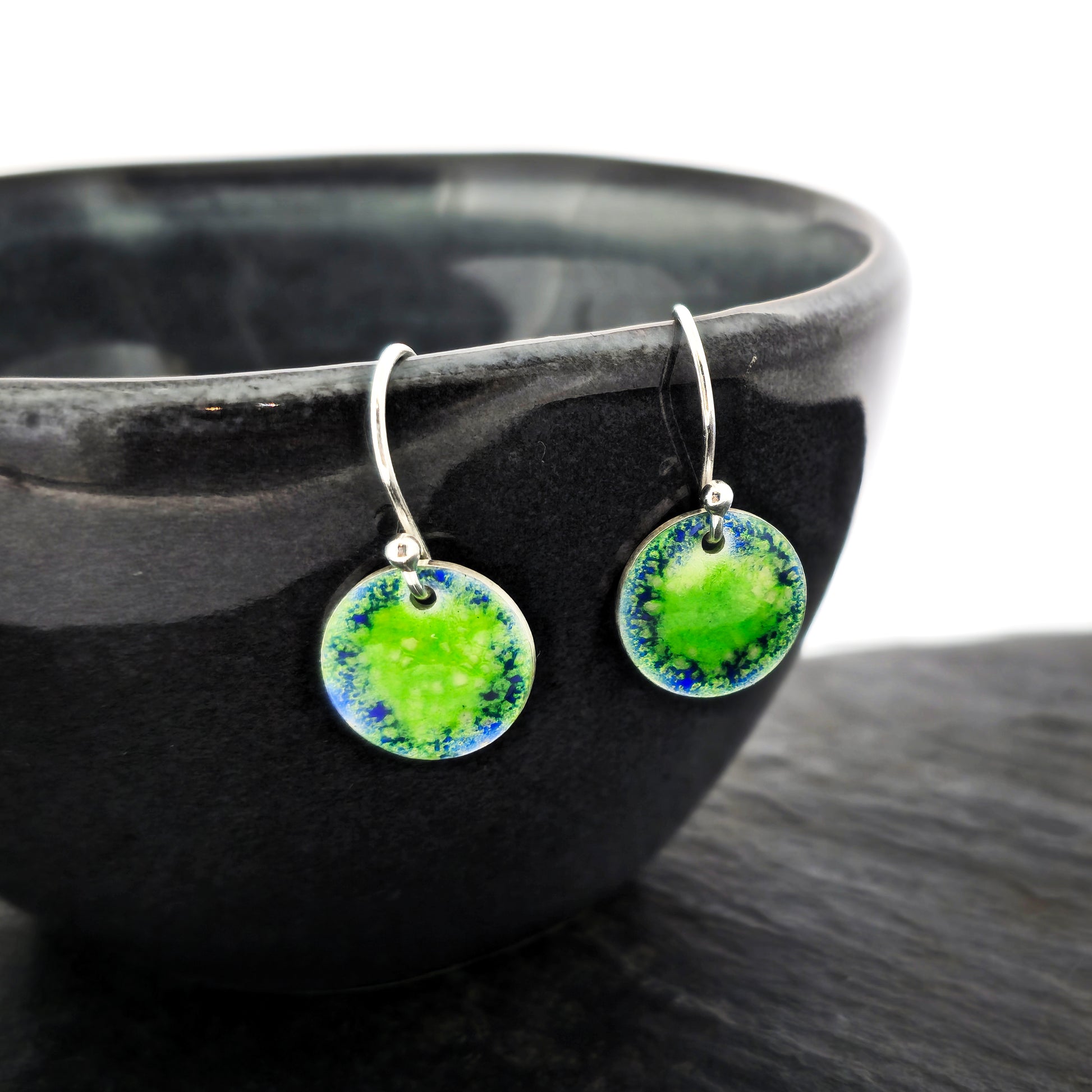 Silver earrings featuring a disc coloured with a pattern of green and blue enamel on silver ear hooks. Pictured on a black bowl.