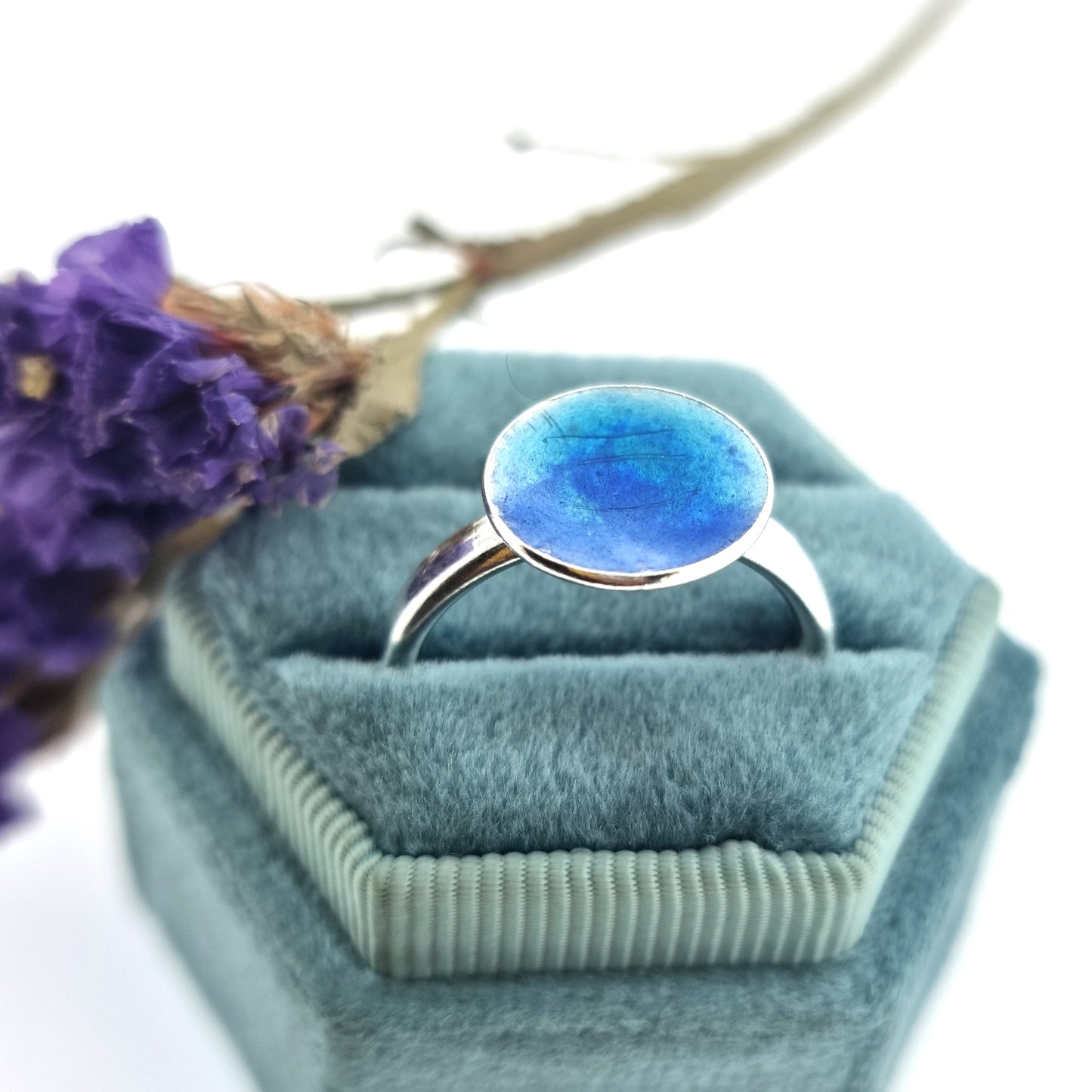 A silver ring band with a round silver top coloured with dark blue and turquoise blue enamel. Pictured in a jewellery box with flowers.
