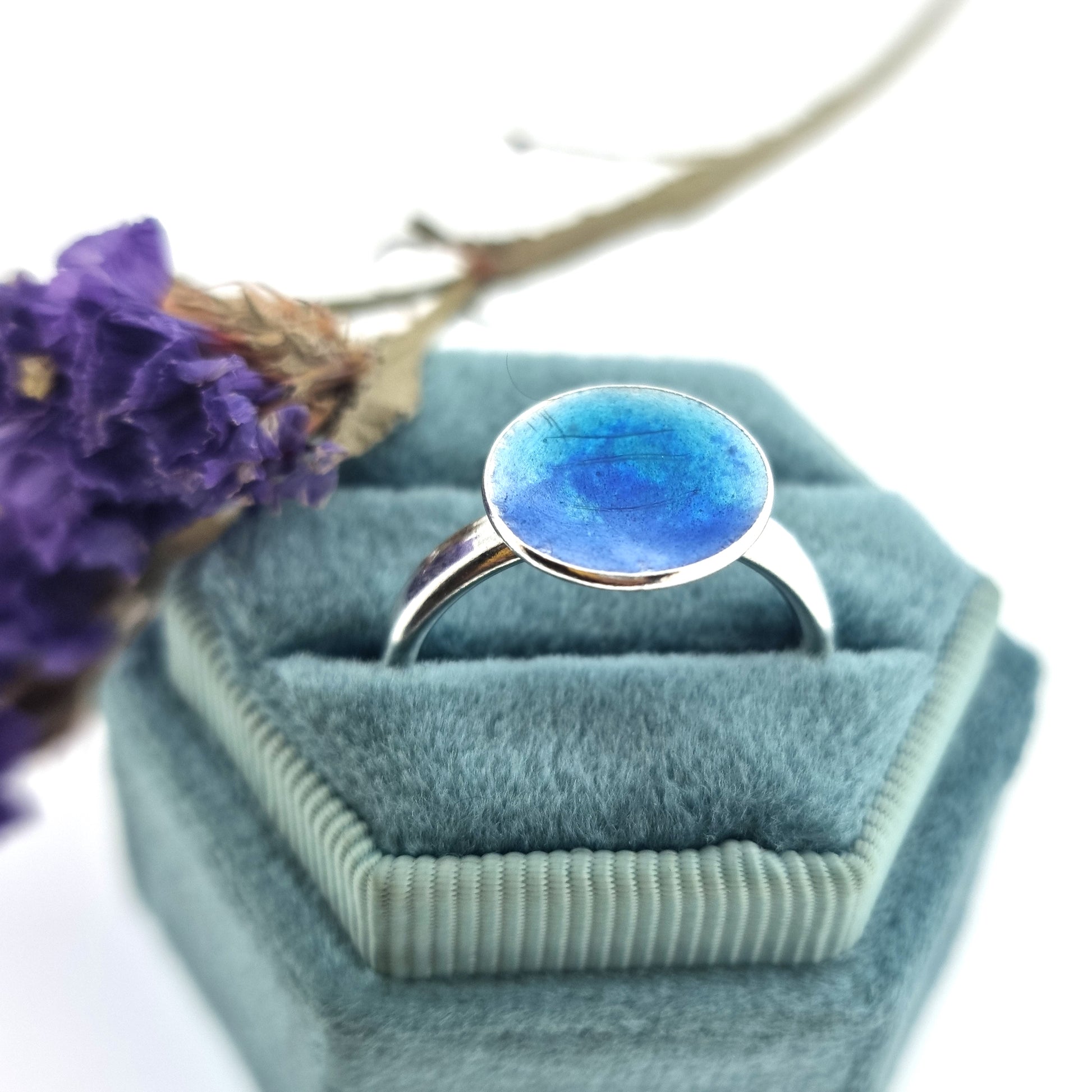 A silver ring band with a round silver top coloured with dark blue and turquoise blue enamel. Pictured in a jewellery box with flowers.