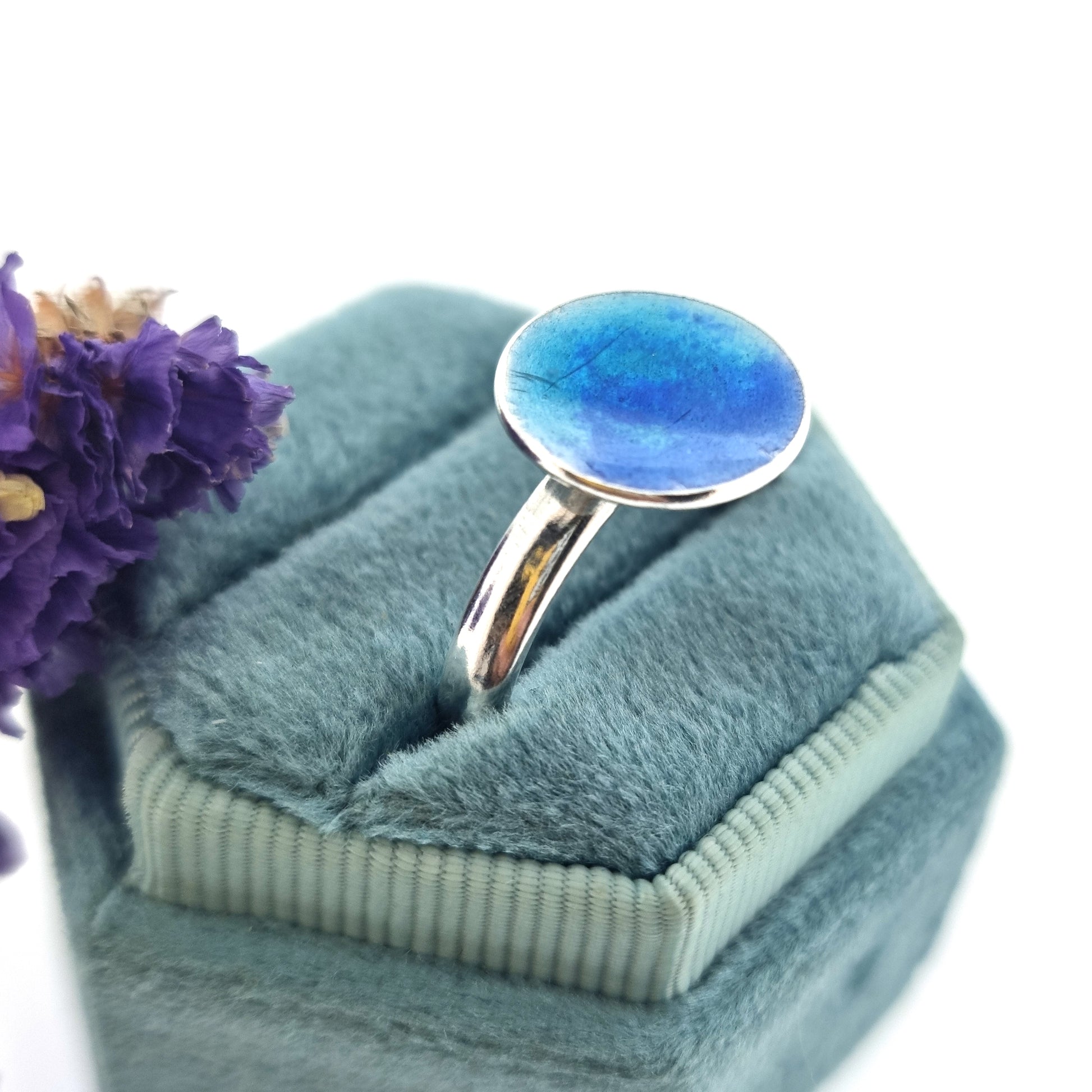 A silver ring band with a round silver top coloured with dark blue and turquoise blue enamel. Pictured n a jewellery box with flowers.