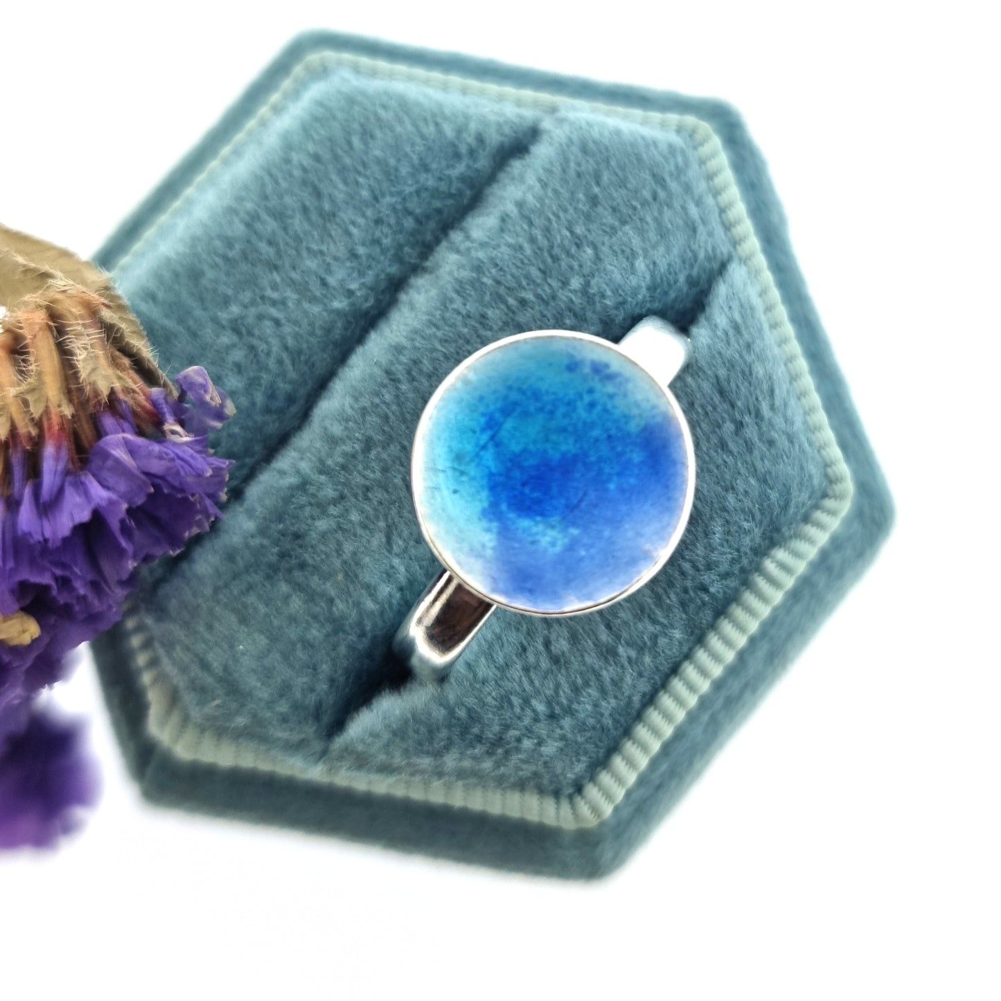 A silver ring band with a round silver top coloured with dark blue and turquoise blue enamel. Pictured n a jewellery box with flowers.
