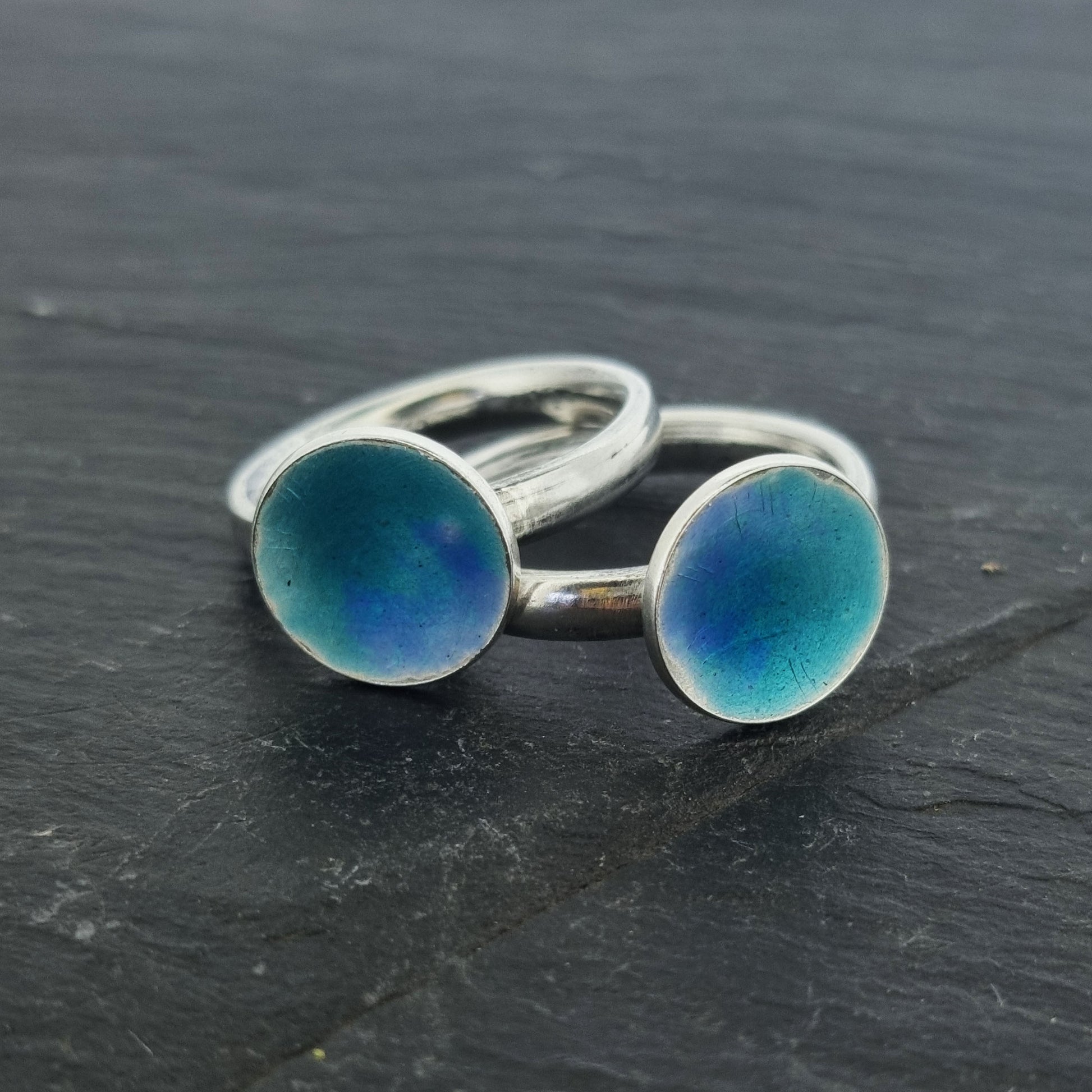 Two silver rings with a silver band with a round silver top coloured with turquoise and turquoise blue enamel. Shown on slate.