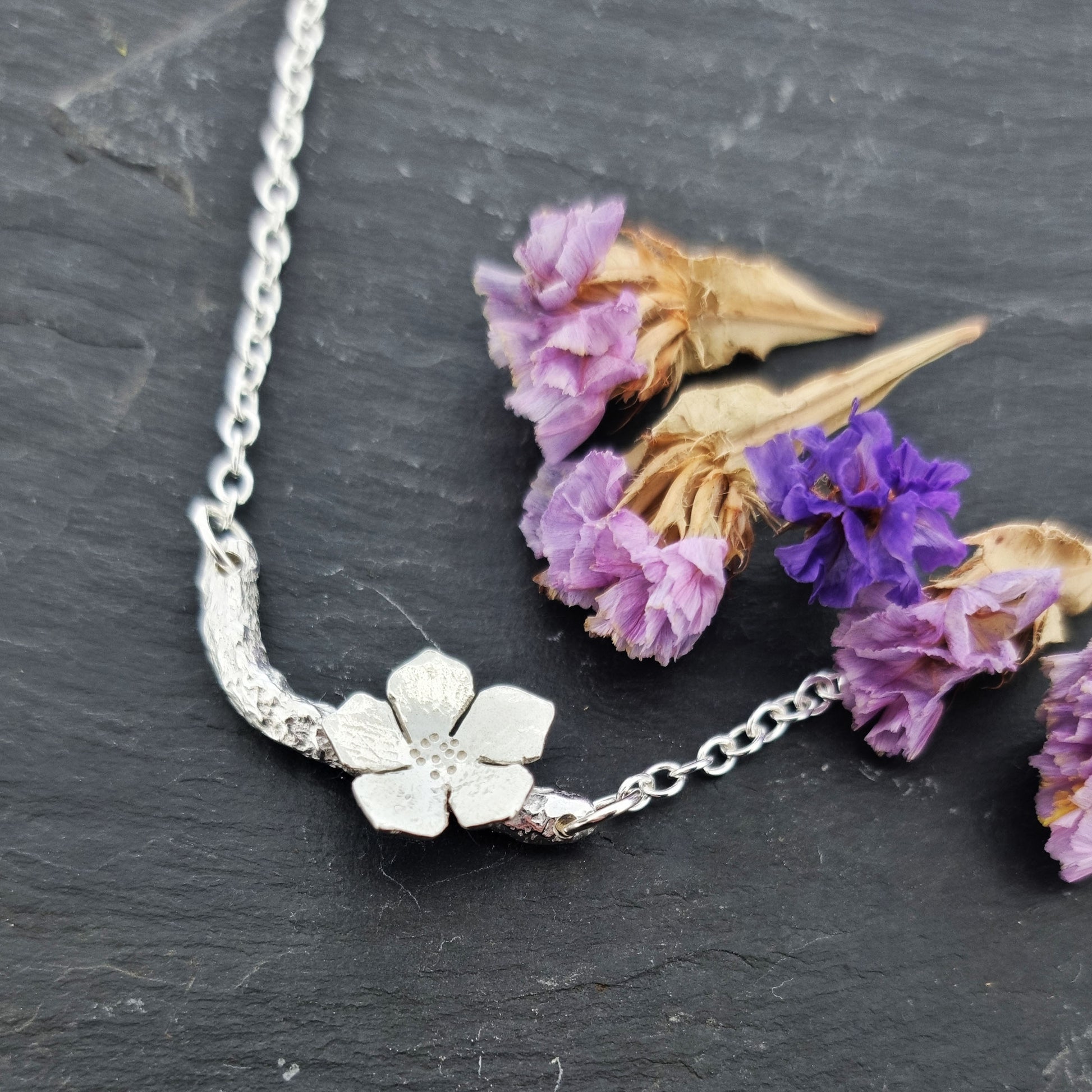 Silver necklace with small branch and a single 5 petal flower on it. Pictured with flowers.
