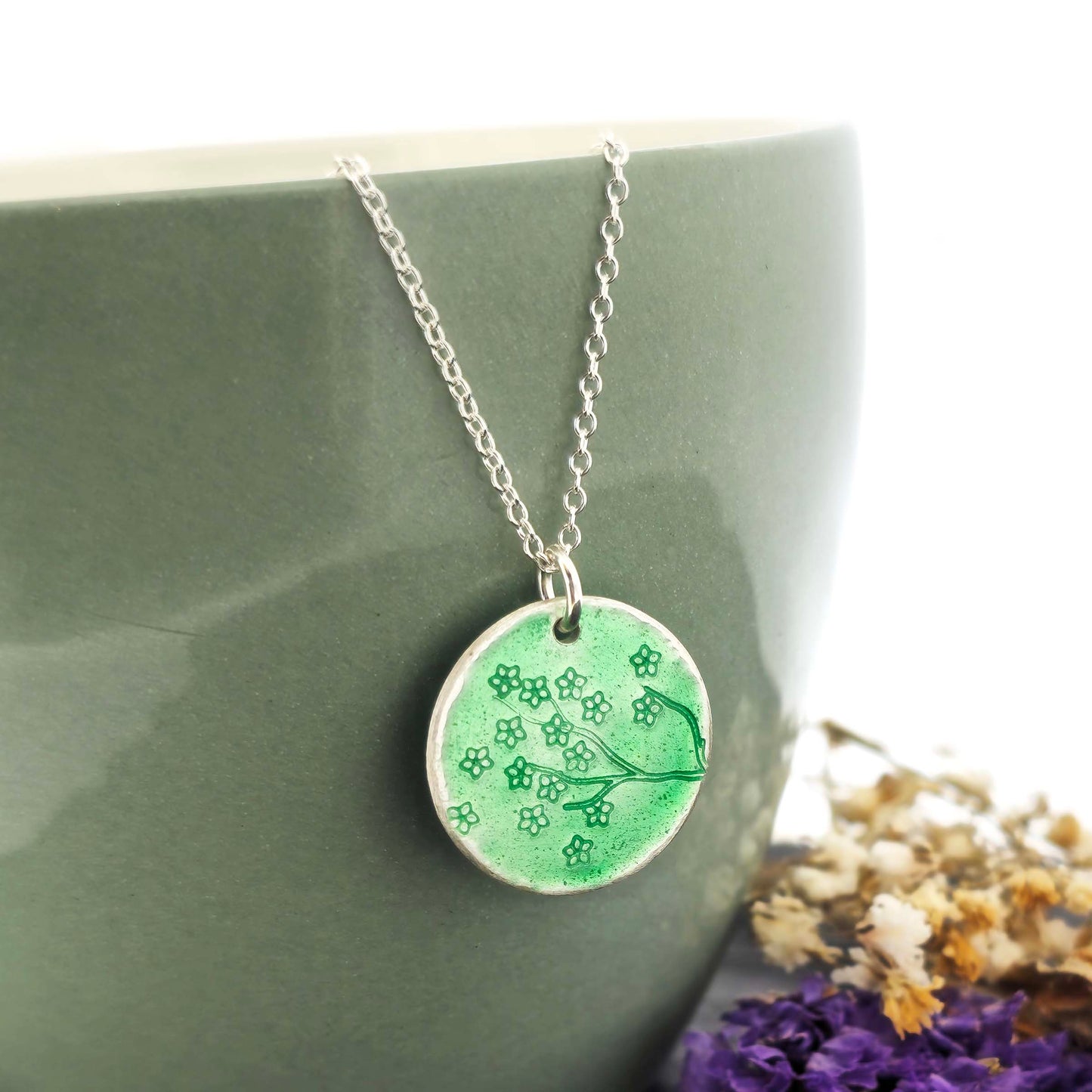 Silver necklace with a round green enamel pendant featuring floral designs on a green surface with flowers in the background.