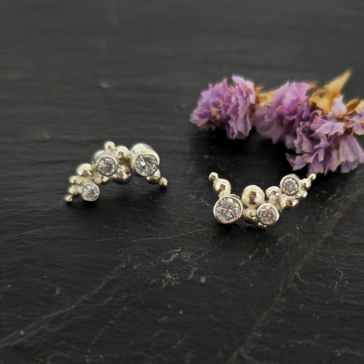 A pair of silver curved earrings made up of balls and gemstones. Pictured on slate with flowers.
