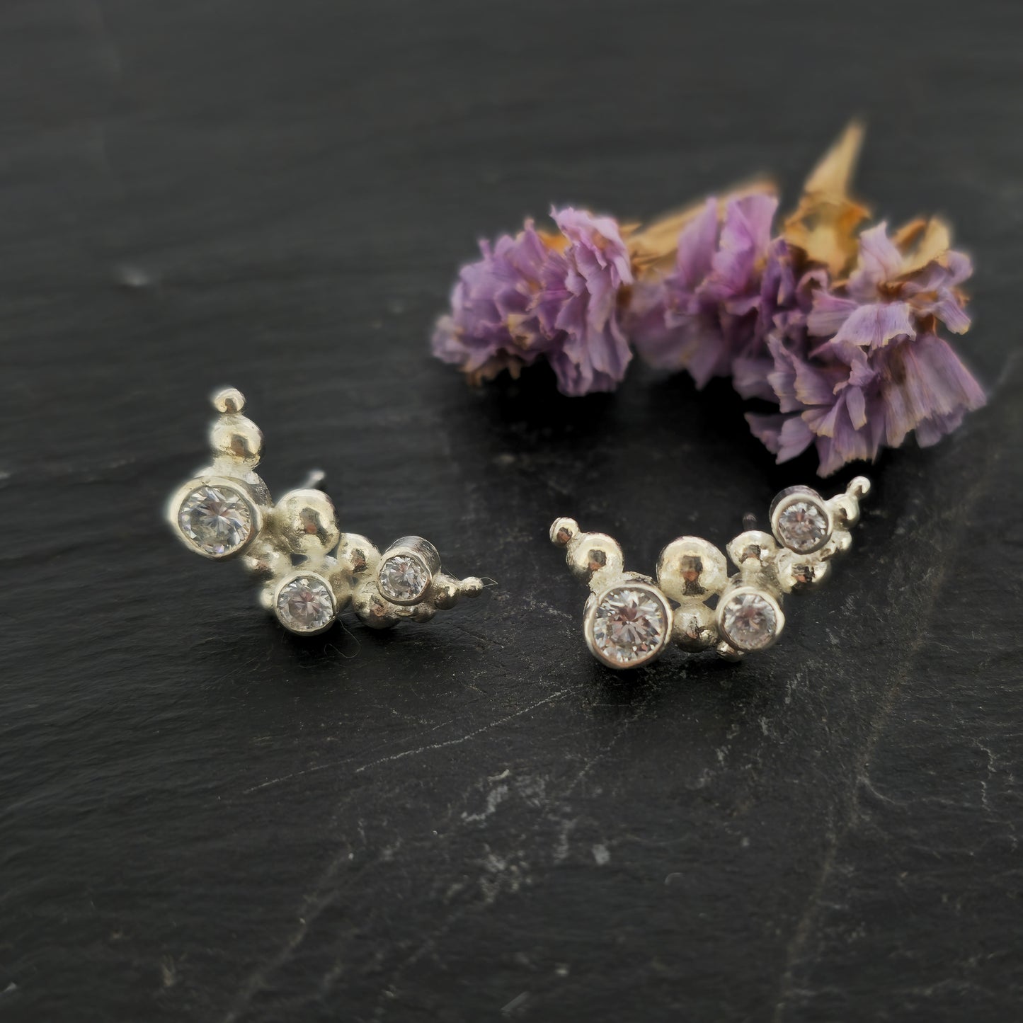 A pair of silver curved earrings made up of balls and gemstones. Pictured on slate with flowers.