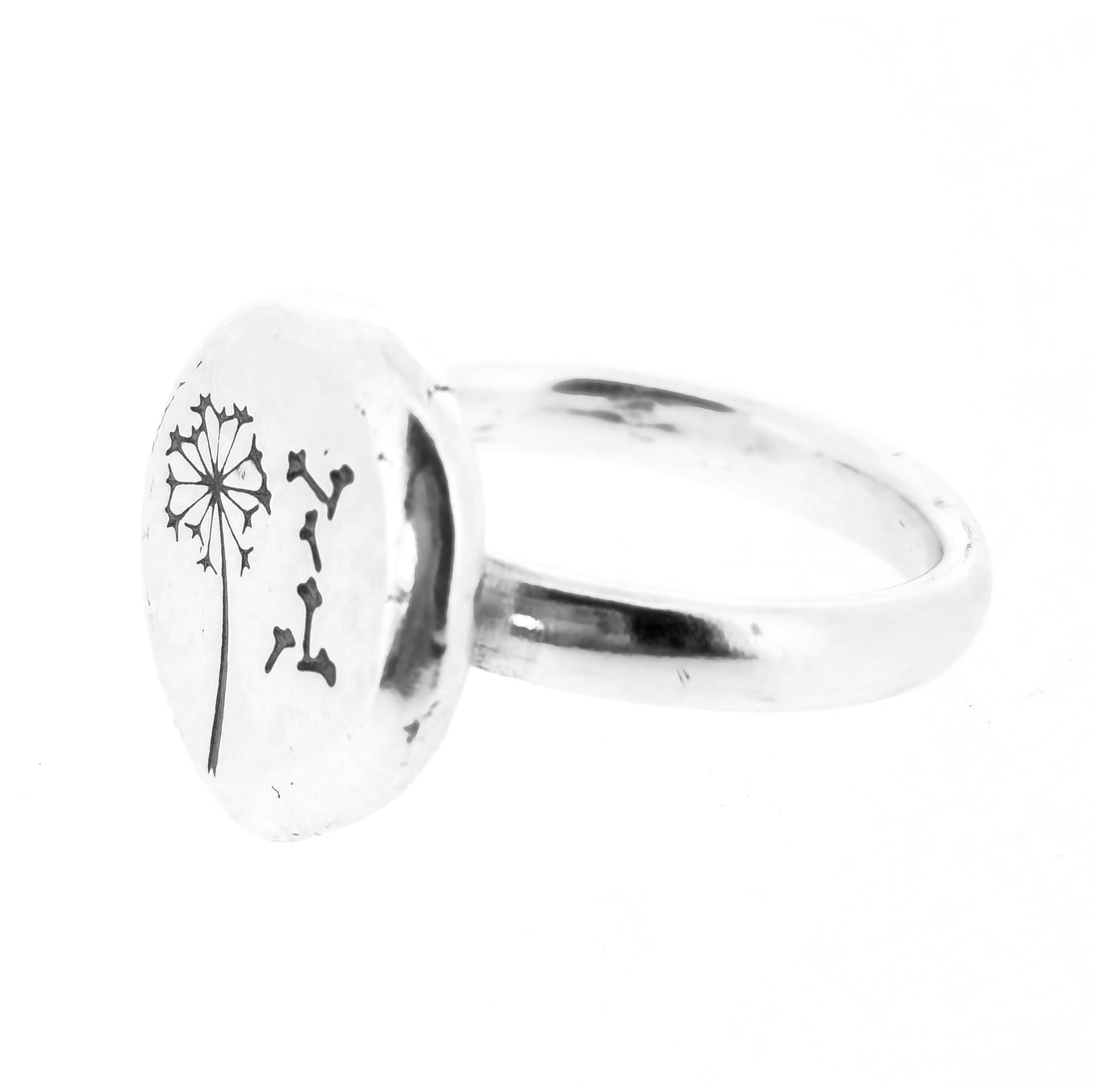 A silver ring with a plain band and a flat pebble top with a dandelion clock and fluff engraved on it.